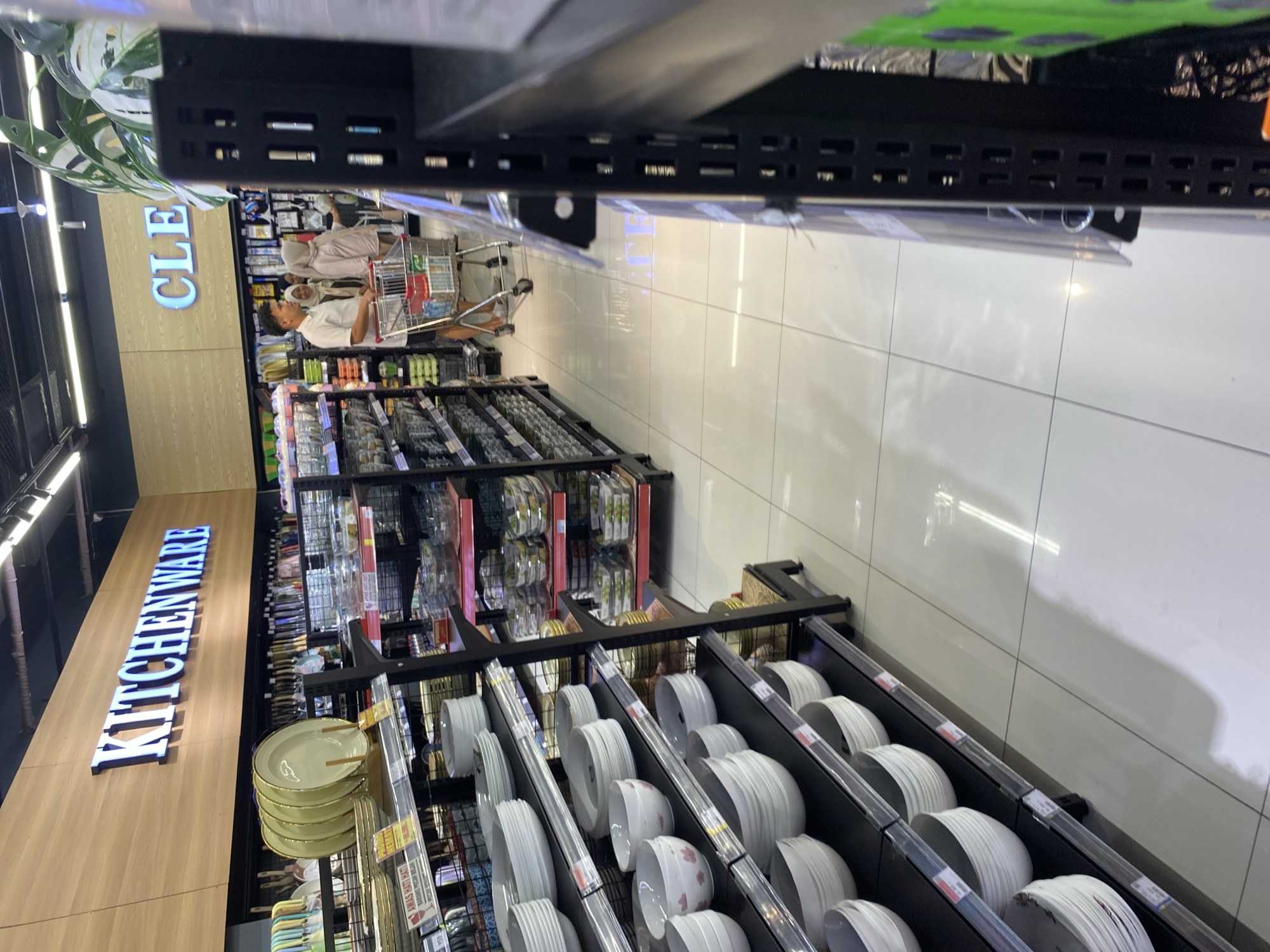 The image shows an aisle in a retail store, likely a department store or hypermarket, specializing in kitchenware and cleaning supplies. In the foreground, shelves are filled with stacks of white ceramic bowls and plates. Further down the aisle, a man and two women are visible, pushing a shopping cart filled with items. The store features illuminated signage that reads "KITCHENWARE" and "CLEAN". The environment appears to be well-lit, with tiled flooring and brightly lit ceiling lights. The overall impression is that of a typical shopping environment in a modern retail establishment. Based on the presence of the "KITCHENWARE" and "CLEAN" signage, and the general layout, this could be a section within a larger store like a department store or hypermarket. The specific location in Banda Aceh, Indonesia cannot be determined from the image alone, as there are no overt geographical markers or local context clues.