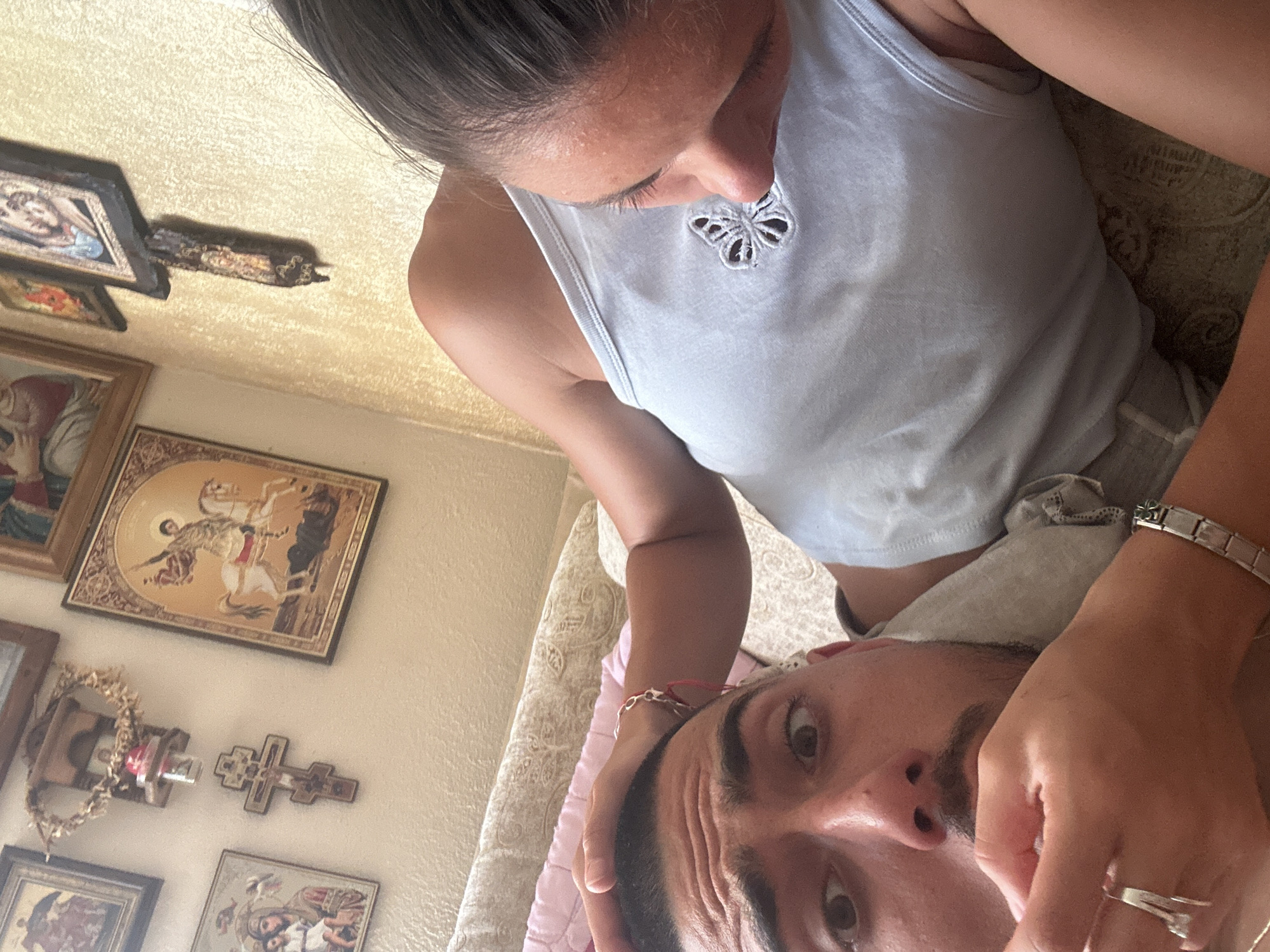 The image is a close-up, overhead shot of a man and woman indoors, possibly in a bedroom in Tankovo, Bulgaria. The man is lying on his back, with his eyes wide open and looking directly at the camera. His hand is near his mouth, and he is wearing a grey shirt. The woman is leaning over him, with her head tilted down, looking towards his chest. She has her hand on his head and is wearing a light blue tank top with a butterfly emblem. In the background, religious icons are visible on the wall, suggesting a home with religious decor. The lighting appears to be natural daylight, creating a warm, intimate atmosphere.