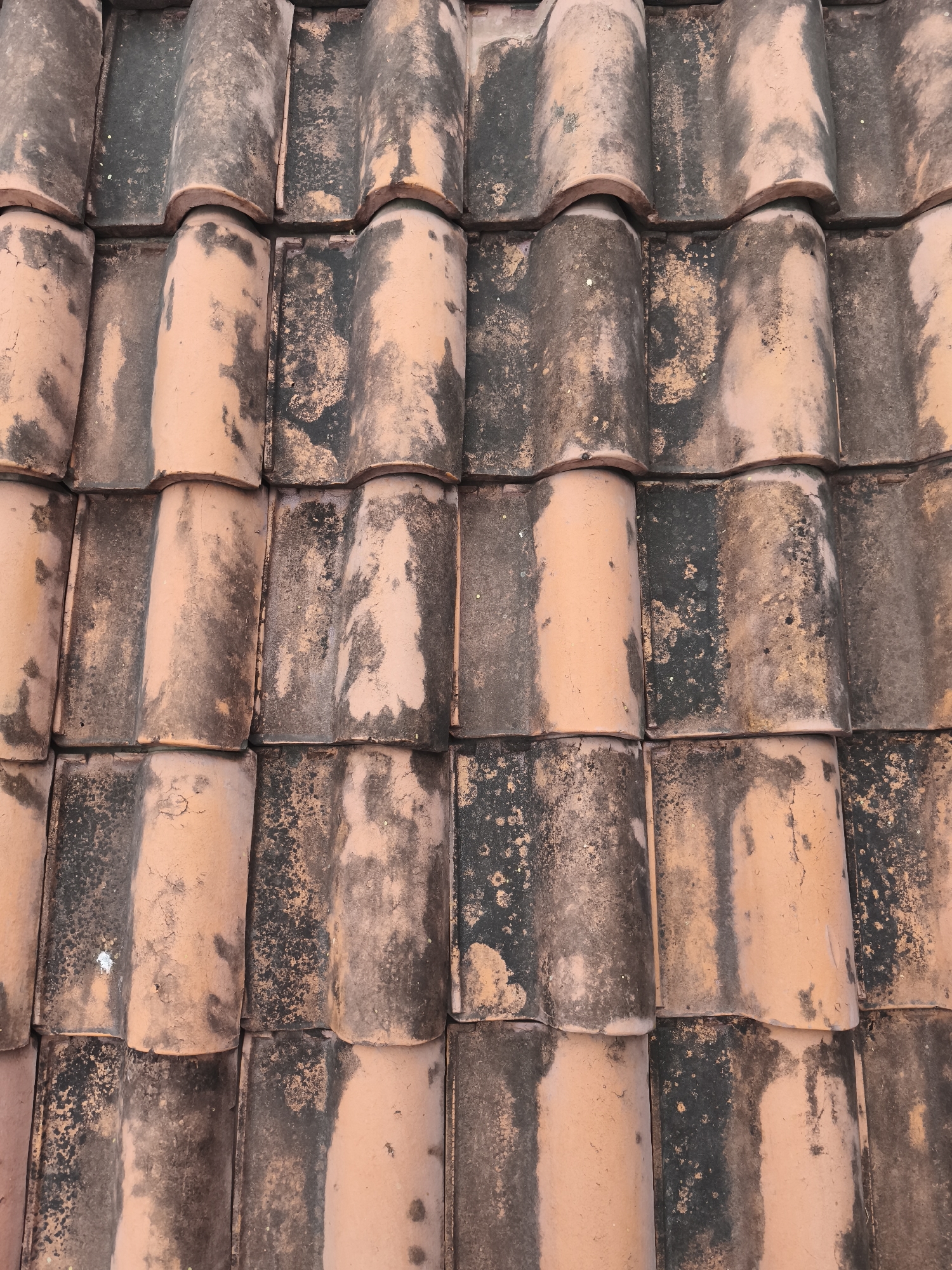 This is a close-up, overhead shot of a terracotta tiled roof, showing the characteristic curved shape of the tiles and their weathered appearance. The tiles exhibit a patchwork of their original reddish-orange hue interspersed with patches of dark discoloration, likely due to algae, moss, or dirt accumulation. The texture appears rough and uneven, with some tiles showing signs of wear and cracking. The lighting suggests it might be daytime, casting subtle shadows that highlight the contours of the tiles. The context provided indicates this scene is located in Yogyakarta, Indonesia.