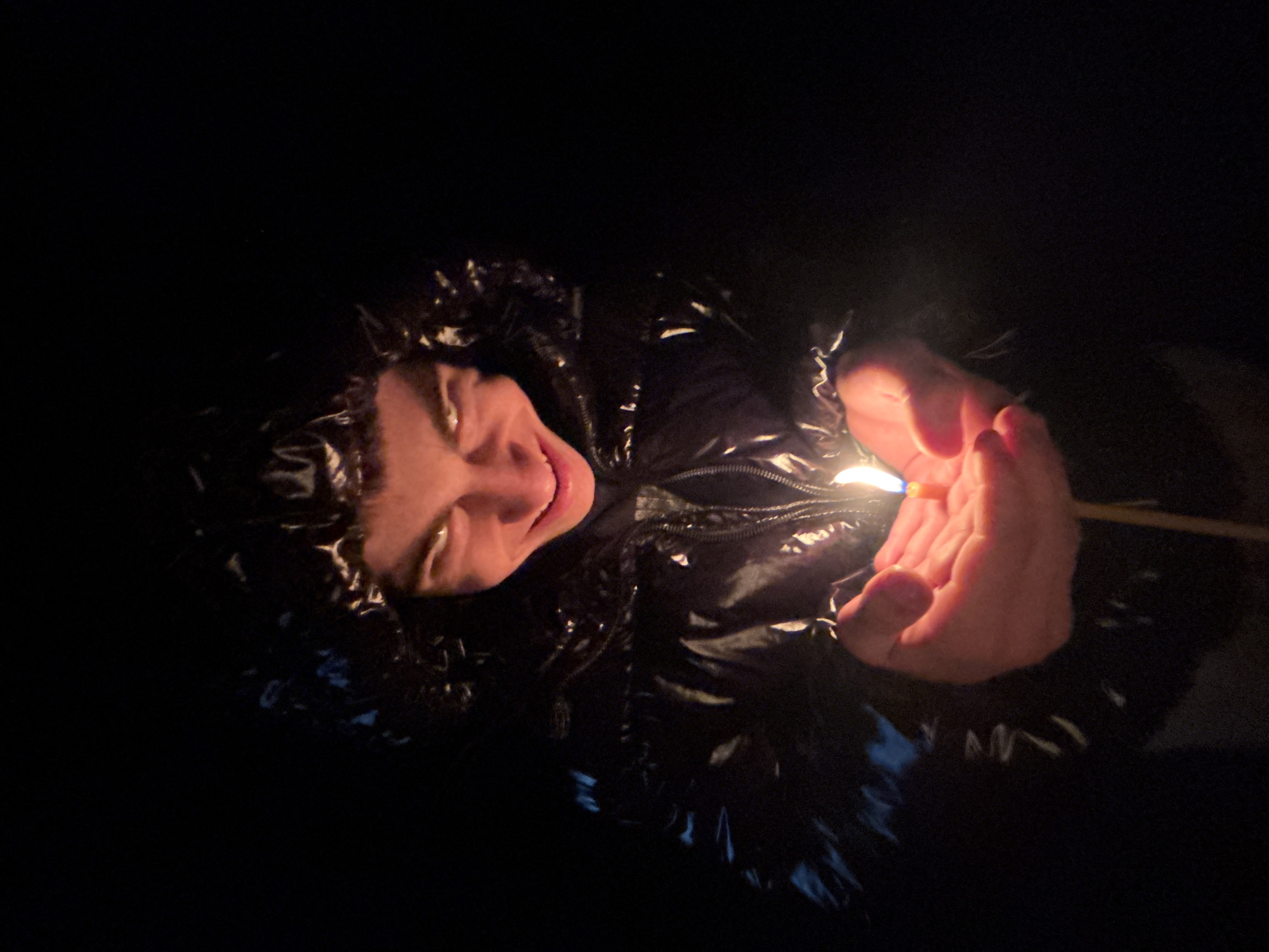 A close-up, high-angle shot shows a person holding a lit match in their cupped hands. The person is wearing a shiny black jacket with a hood. The light from the match illuminates the person's face, showing a smile. The background is dark. The location is not discernible from the image.
