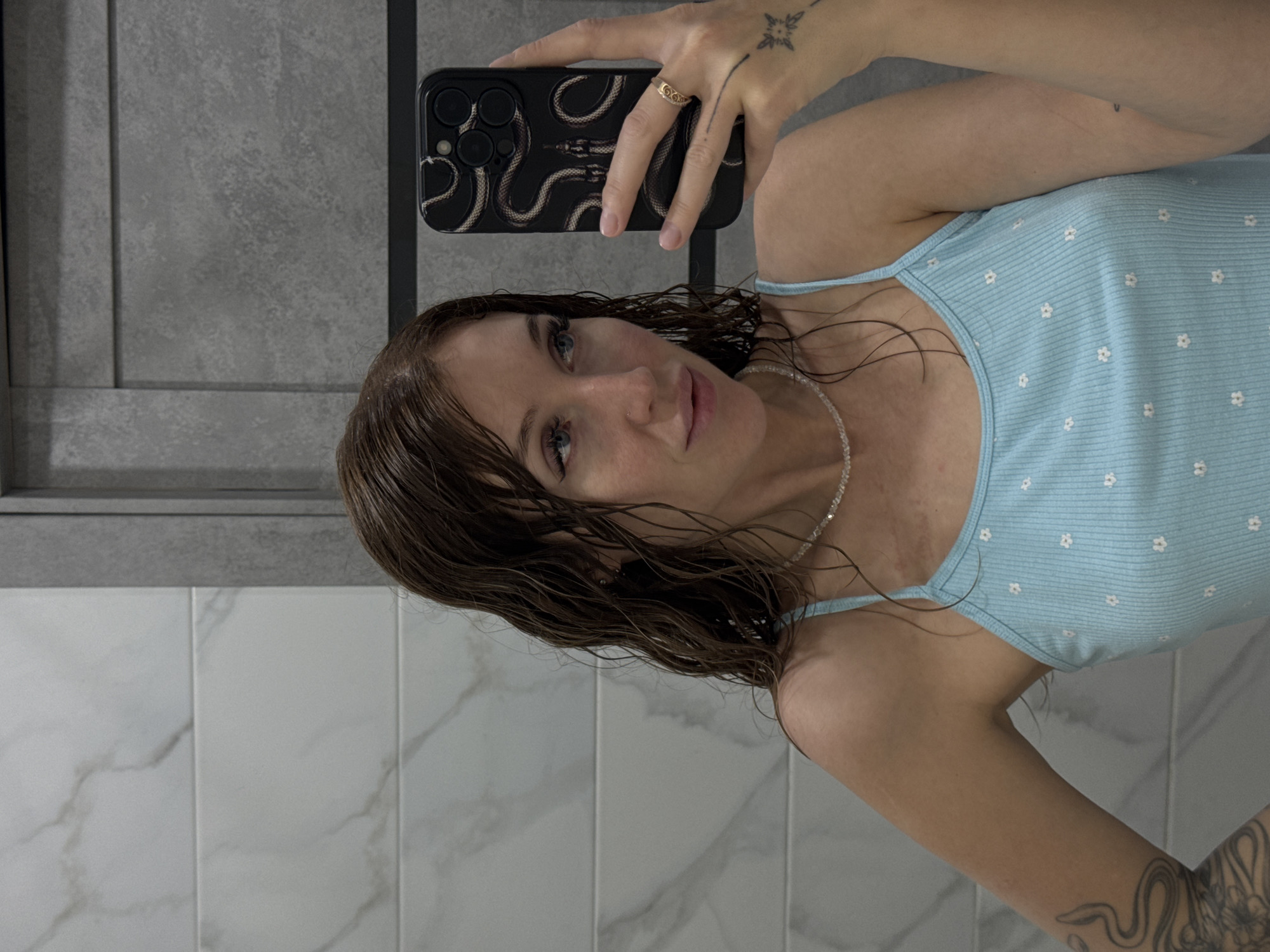 A woman with wet, brown hair and blue eyes takes a selfie in a bathroom. She is wearing a light blue tank top with a floral pattern and a beaded necklace. She has tattoos on her arm and hand, and a ring on her finger. The bathroom has white tiled walls with grey grout, and a grey tiled section. The photo appears to be taken in Chaikovsky, Russia.