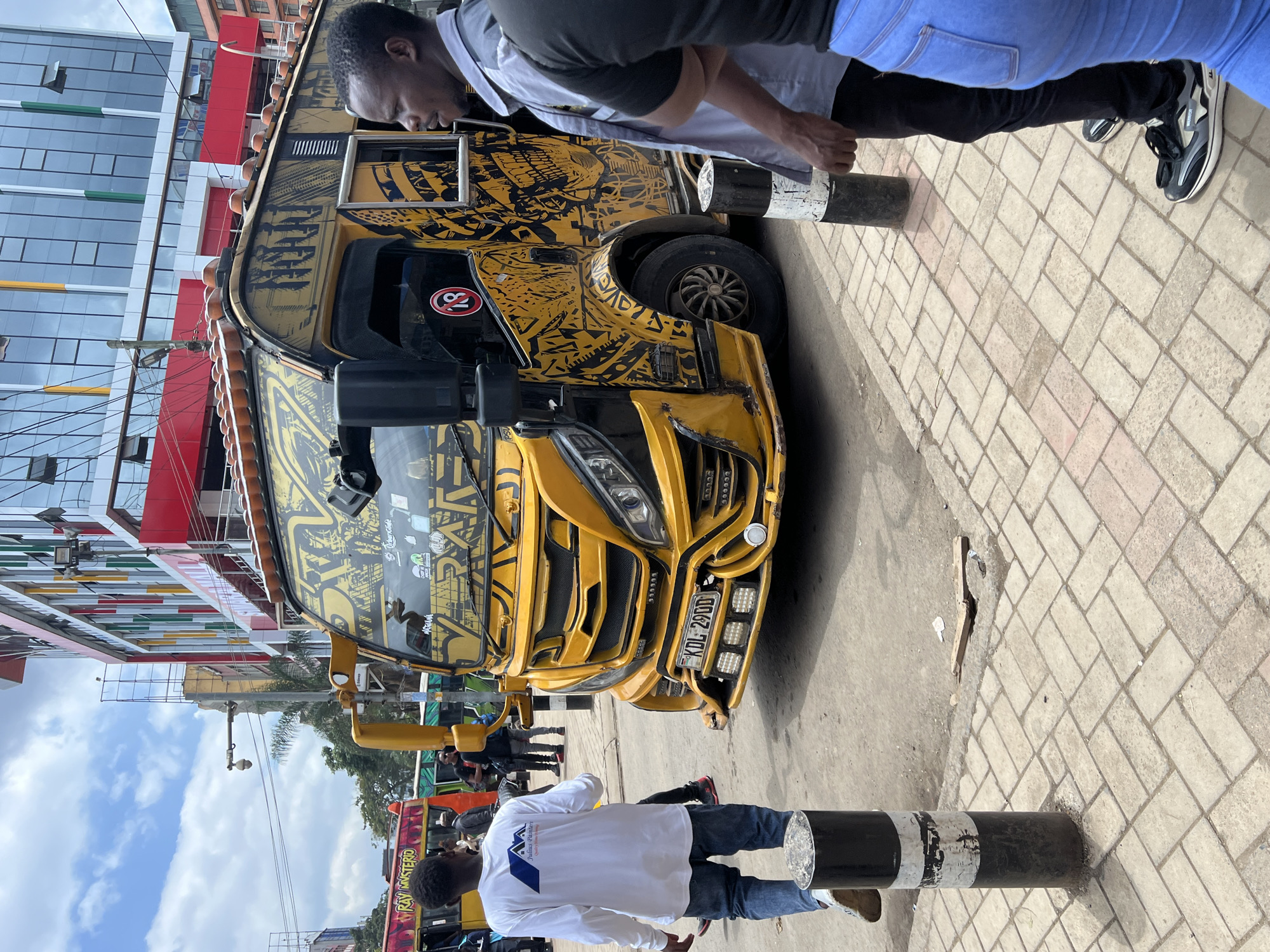 The image captures a vibrant daytime urban street scene, likely in Nairobi, Kenya, under a partially cloudy sky. The central subject is a highly customized yellow and black matatu (public service vehicle), which is parked on the left side of a paved road next to a sidewalk. The matatu is adorned with intricate black graphic patterns, numerous additional lights on its front bumper, and displays the license plate "KDL 290U." A prominent red circle with the number "18" is visible on its side, along with the word "FURY" painted near its roofline.
Two men are visible in the foreground and midground. In the foreground, a man is seen from behind, wearing a white long-sleeved shirt and blue jeans, standing next to a black and white striped bollard. His shirt features a blue mountain-like logo and the text "DALUAS PAINTERS Quality & Style." Another man, partially visible in the upper right, wears a dark t-shirt over a white collared shirt and black trousers. He stands near the matatu, appearing to interact with it or wait, also beside a black and white bollard.
In the background, modern multi-story buildings with glass facades and colorful horizontal accents in red, green, and yellow hues line the street. Further down the road, another matatu is partially visible, bearing the text "RAY MYSTERO." The ground consists of a light-colored, herringbone-patterned paved sidewalk and a darker asphalt road. The bright lighting suggests it is mid-day, and the overall scene reflects the distinctive atmosphere of a bustling Kenyan city.
