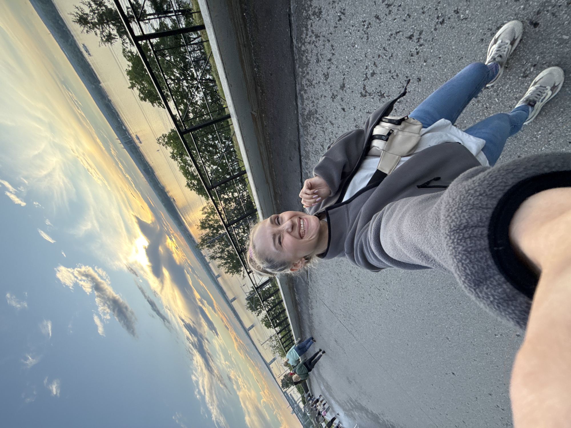 The image captures a selfie taken by a young Caucasian woman with blonde hair pulled back in a bun, smiling widely. She is wearing a grey fleece jacket over a white top, blue jeans, and grey sneakers. A small beige fanny pack is worn across her chest. The background reveals a panoramic view of the Kama River in Perm, Russia, at sunset. The sky is a canvas of vibrant orange, yellow, and blue hues, with scattered clouds. A long bridge, likely the Permsky Bridge, spans the river in the distance, and several boats are visible on the water. A black metal fence lines the embankment where the woman is standing, separating her from the river and its tree-lined opposite bank. In the middle ground, other people are walking along the paved walkway. The overall impression is a candid moment of enjoyment during a scenic evening in Perm.