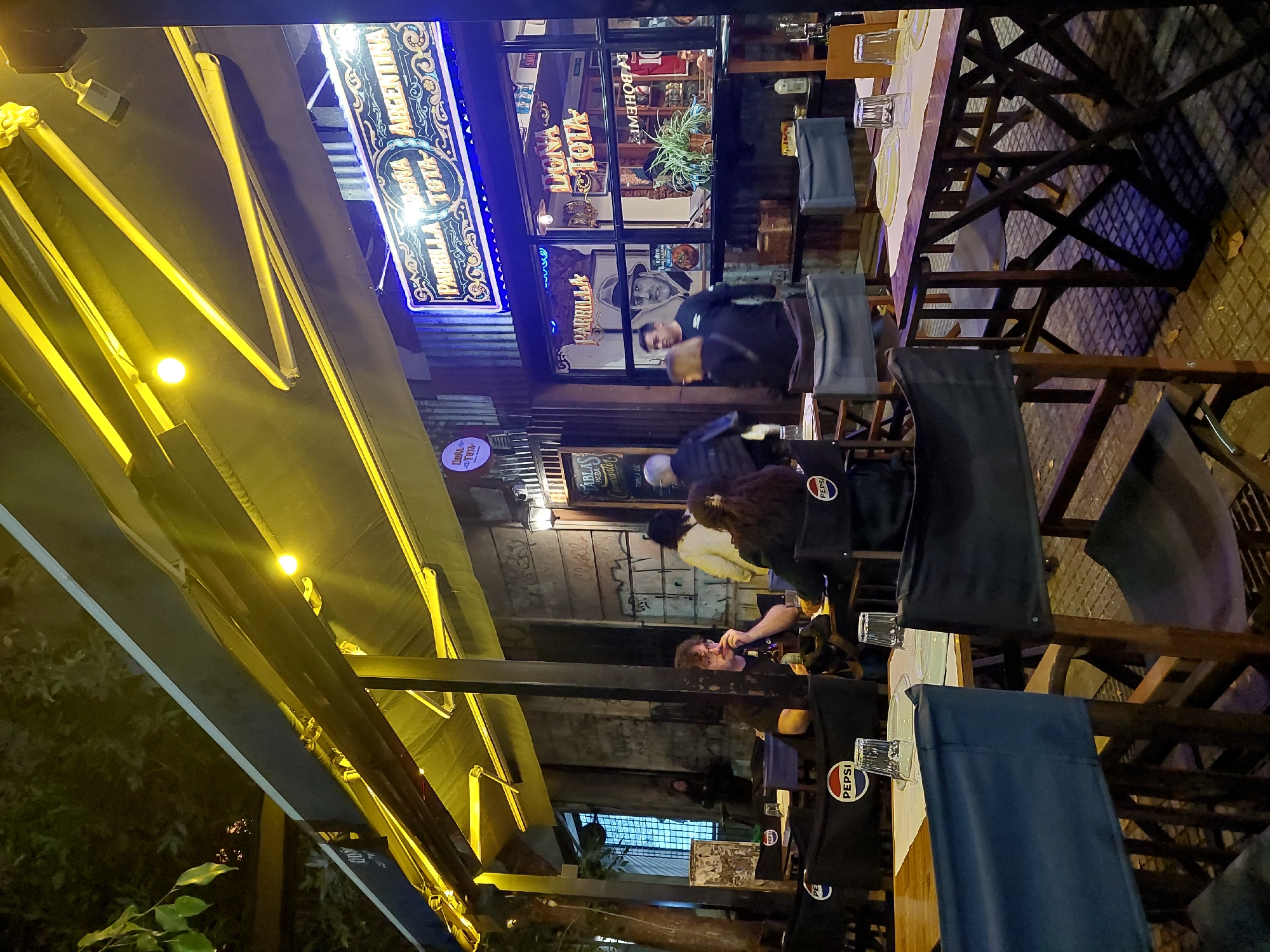 Here is a detailed description of the image:
**Scene:** The image depicts the outdoor seating area of "Parrilla Dona Tota" restaurant, which is located in Buenos Aires, Argentina, as seen by signage indicating "Parrilla Dona Tota Argentina" and the "La Bohemia" establishment next door.
**People:** Several people are seated at tables, partaking in food and drinks. One person is walking near the restaurant windows. Some are partially obscured by chairs and table coverings.
**Objects:** The scene is comprised of outdoor dining furniture, including wooden tables and chairs covered with blue fabric displaying "Pepsi" logos. The tables are set with glasses and plates. Overhead are lights and a fabric awning covering the outdoor dining area. The restaurant's illuminated signage is visible.
**Environment:** The scene is an outdoor restaurant at night. The lighting creates a somewhat dark atmosphere, with areas illuminated by lights and the restaurant's signage.