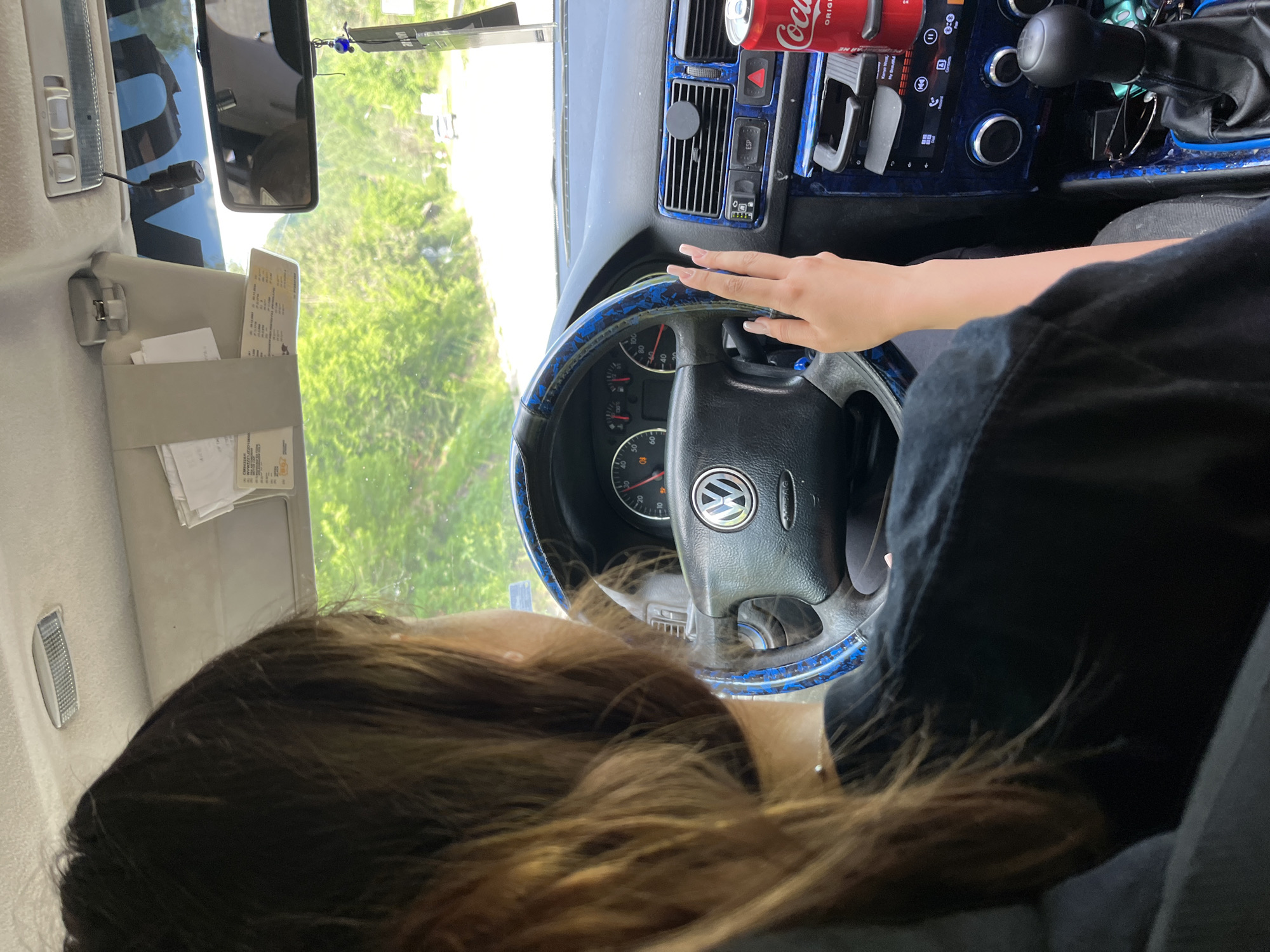 The image depicts the interior of a car, viewed from the passenger seat, with a person's head and shoulders visible from the back. The driver, a young female with long brown hair, is behind the steering wheel, her left hand resting on it and her right hand extended slightly forward. The car's dashboard is visible, featuring a Volkswagen logo on the steering wheel, a speedometer, and various controls. A can of Coca-Cola is placed in a cup holder on the right side of the dashboard. Outside the car, a road is visible, lined with green trees, suggesting a rural or suburban setting. The location is identified as Vlahovo, Bulgaria.
