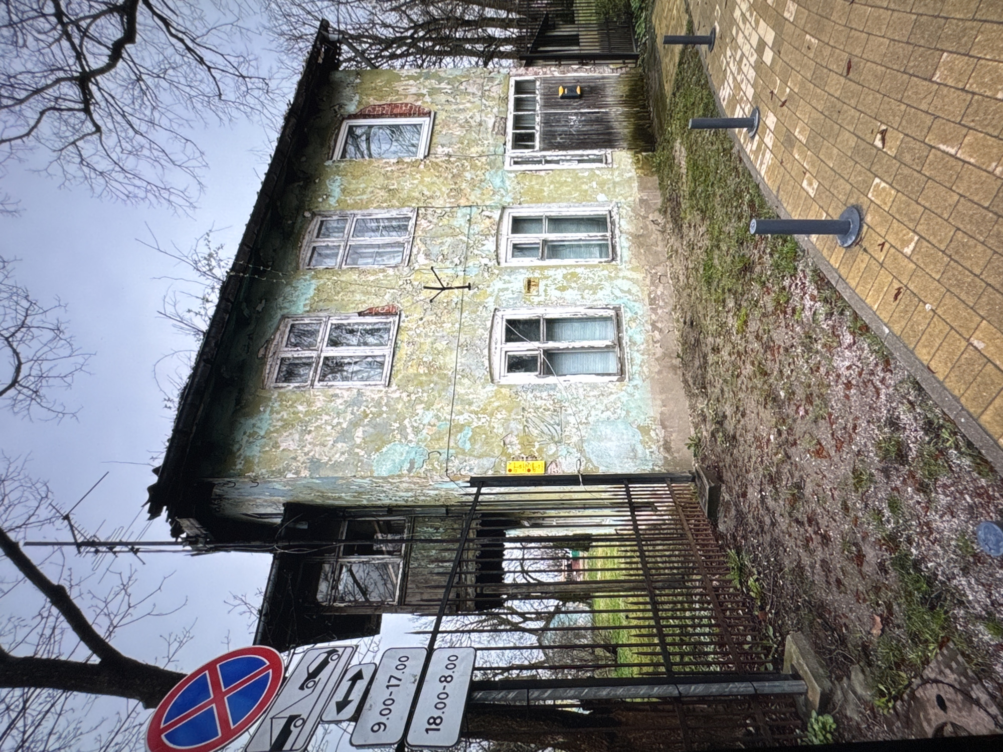 The image depicts a weathered, three-story building in Новинки, Russia. The building's facade exhibits peeling paint and several window openings, some of which are boarded up. A metal fence with multiple road signs, including a "No Parking" sign and time restrictions (9.00-17.00 and 18.00-8.00), stands in front of the building. A brick sidewalk runs along the side of the building, marked with gray cylindrical bollards. The sky is overcast, and bare tree branches are visible in the background. The surrounding area appears to be a mix of vegetation and unpaved ground.