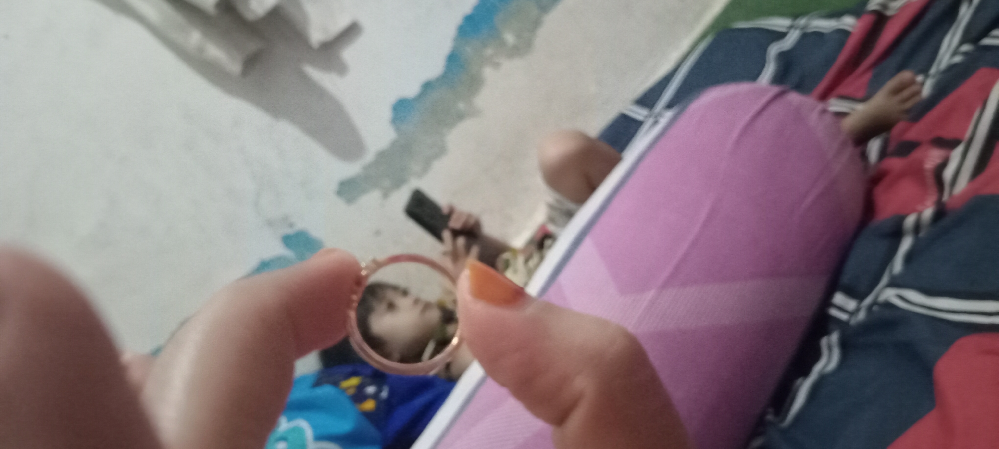 A hand, with orange nail polish, holds a small circular mirror. The mirror reflects a child looking at a smartphone. The child is wearing a light-colored shirt. In the background, a light-colored wall is visible with blue and green paint splotches. The child is seated on a surface covered with a pink pillow and a blue and red striped blanket. The lower portion of the image shows a child's foot. The scene appears to be indoors.