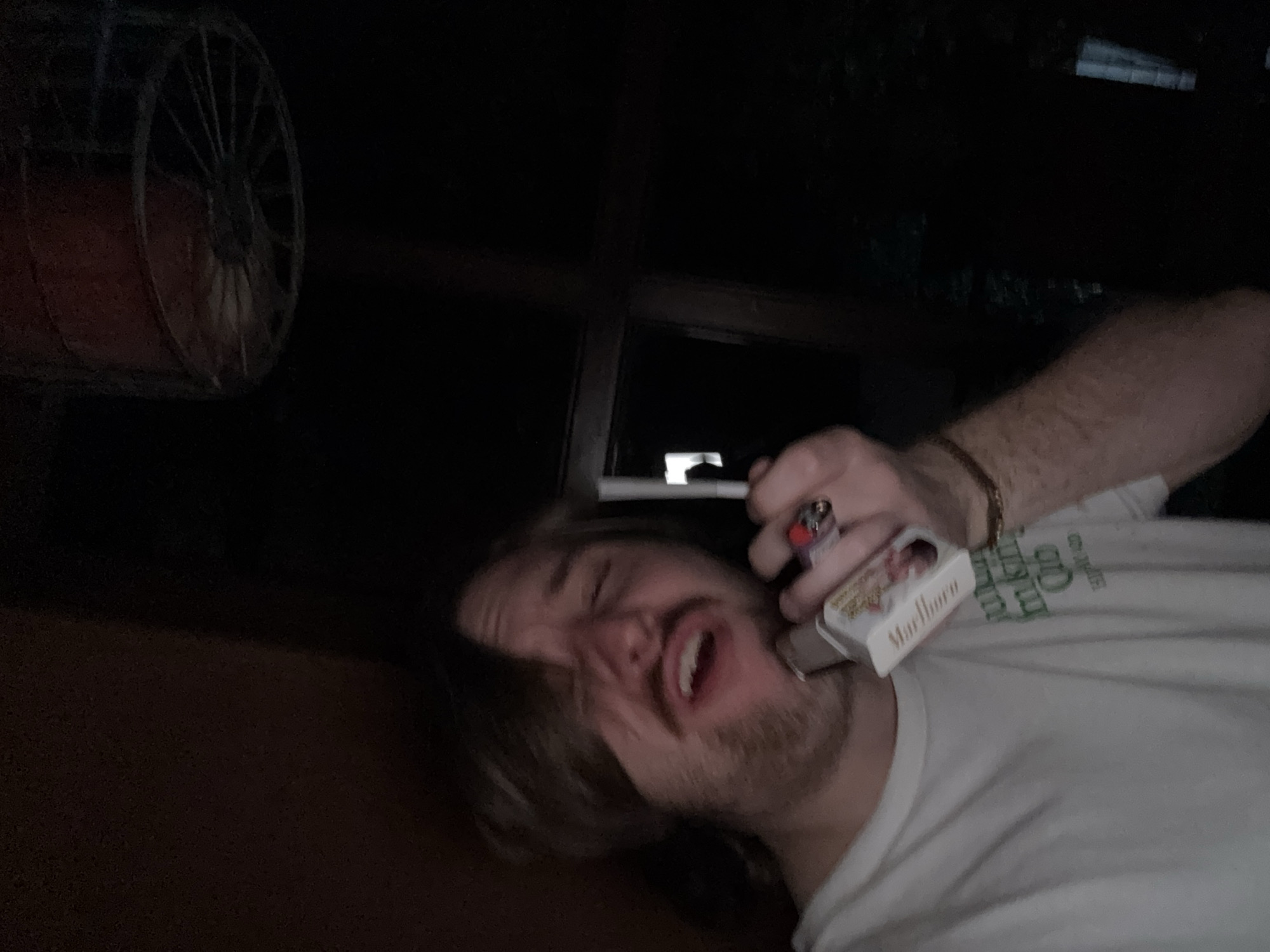 A man with long hair and a beard holds a cigarette to his lips, a pack of Marlboro cigarettes against his face, and a lighter in his hand. He is wearing a white t-shirt and a bracelet on his wrist. A decorative wheel is visible in the background. The image was taken at night. The location is Cañada de Madero, Mexico.