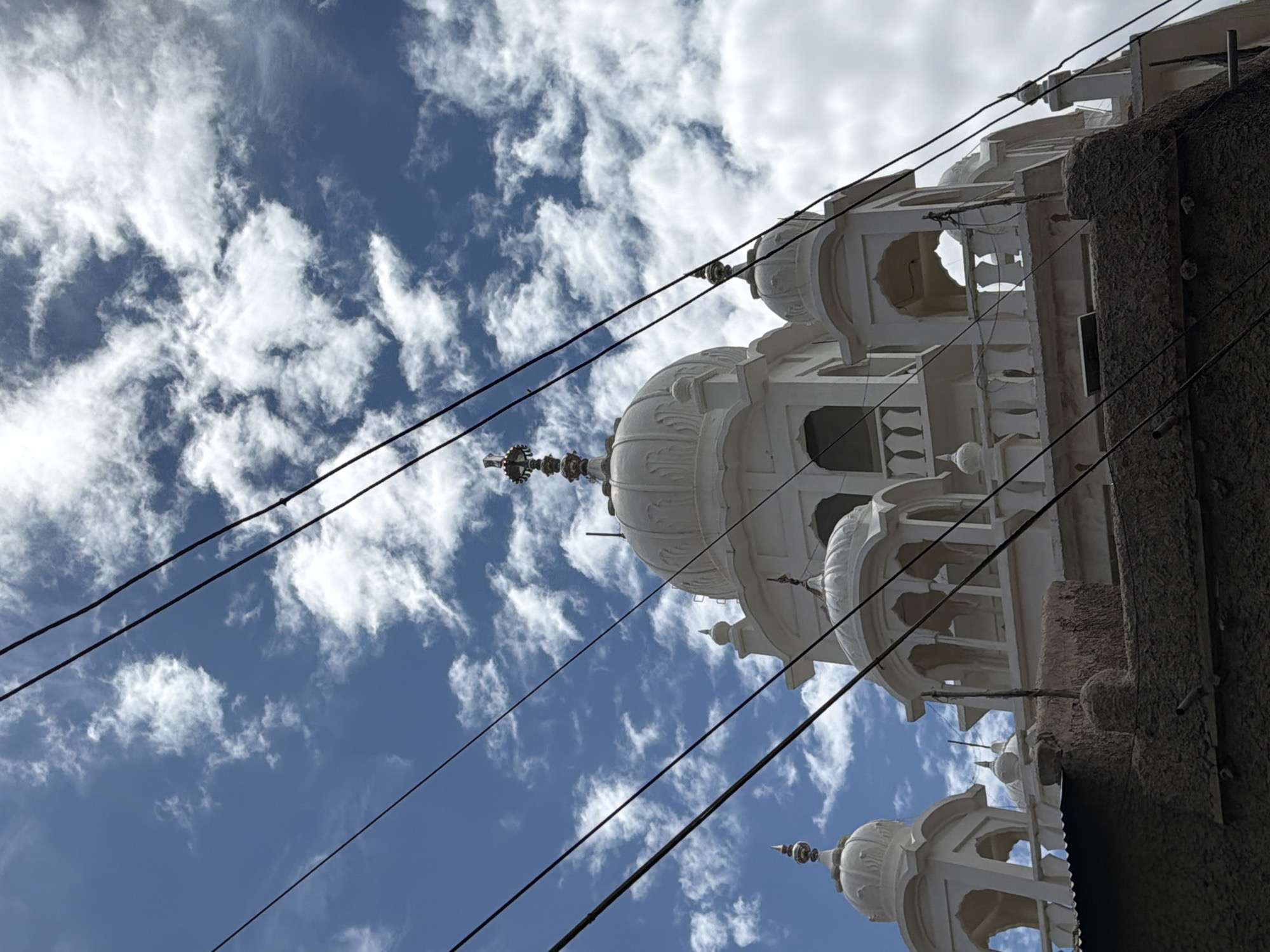 The image captures a low-angle view of a white gurdwara, likely in Leh, India, set against a backdrop of a bright blue sky dotted with fluffy white clouds. Several black electrical wires crisscross the foreground, obscuring parts of the building's upper sections. The gurdwara features multiple domes, arches, and decorative railings, typical of Sikh architecture. In the lower part of the frame, a rough, dark-colored wall or rooftop is visible, providing a contrast to the pristine white structure above. The scene is illuminated by daylight, suggesting it was taken during the daytime. There are no people visible in the image.