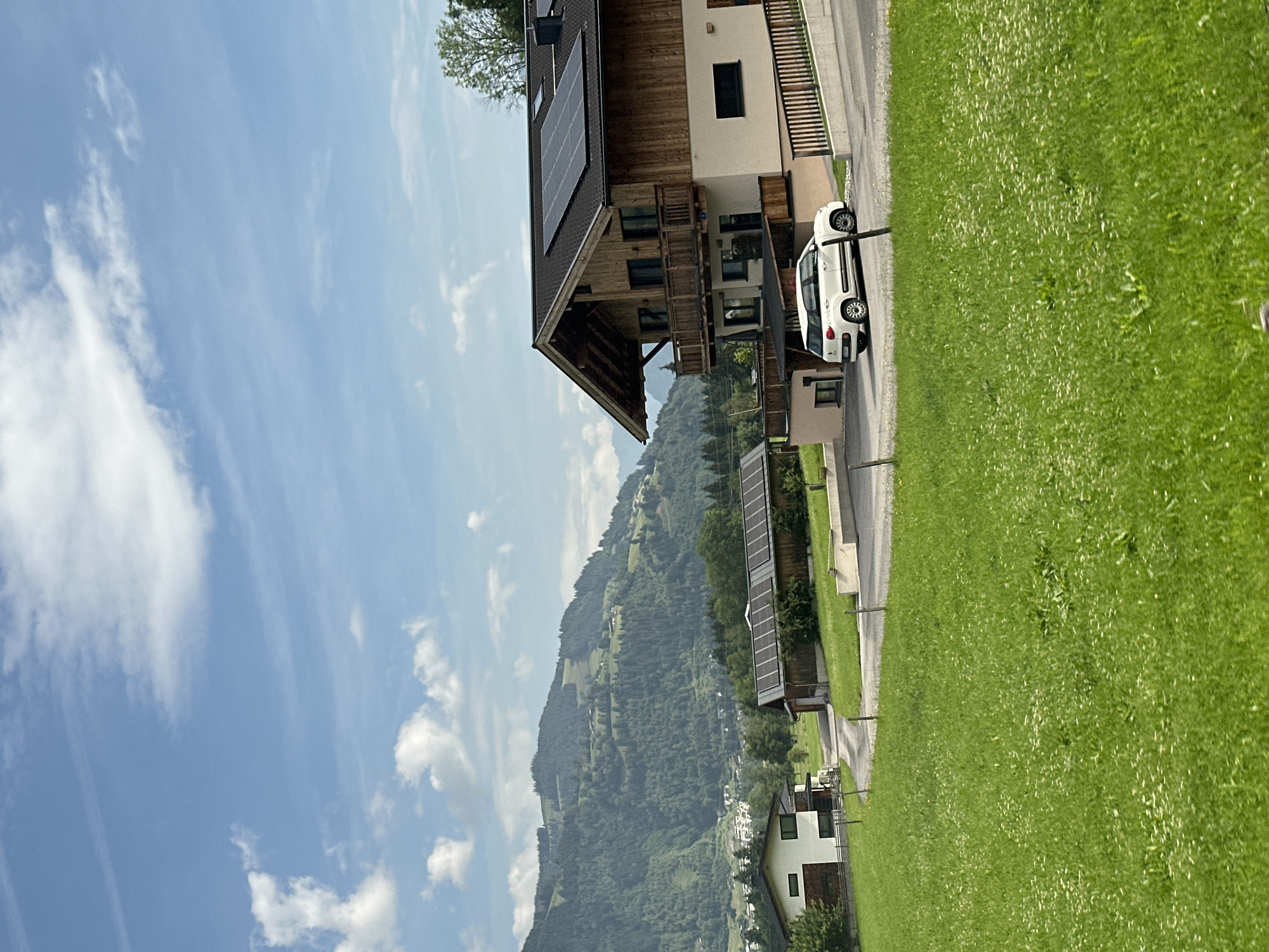 The image depicts a landscape scene in Kaprun, Austria, oriented vertically. The foreground is dominated by a vibrant green lawn that slopes gently upwards. Adjacent to the lawn is a paved pathway and a building with a wooden facade, solar panels on its roof, and a balcony. A small white car is parked beside the building.
In the background, a mountain range covered in lush green trees stretches across the frame. The sky is a bright blue, dotted with scattered white clouds, suggesting it is daytime with fair weather. The overall atmosphere is serene and idyllic, typical of a rural mountainous region. The presence of solar panels on the building suggests an emphasis on sustainable energy.
There is no visible text in the image. The scene appears to be a snapshot of a peaceful residential area nestled in the Austrian Alps.