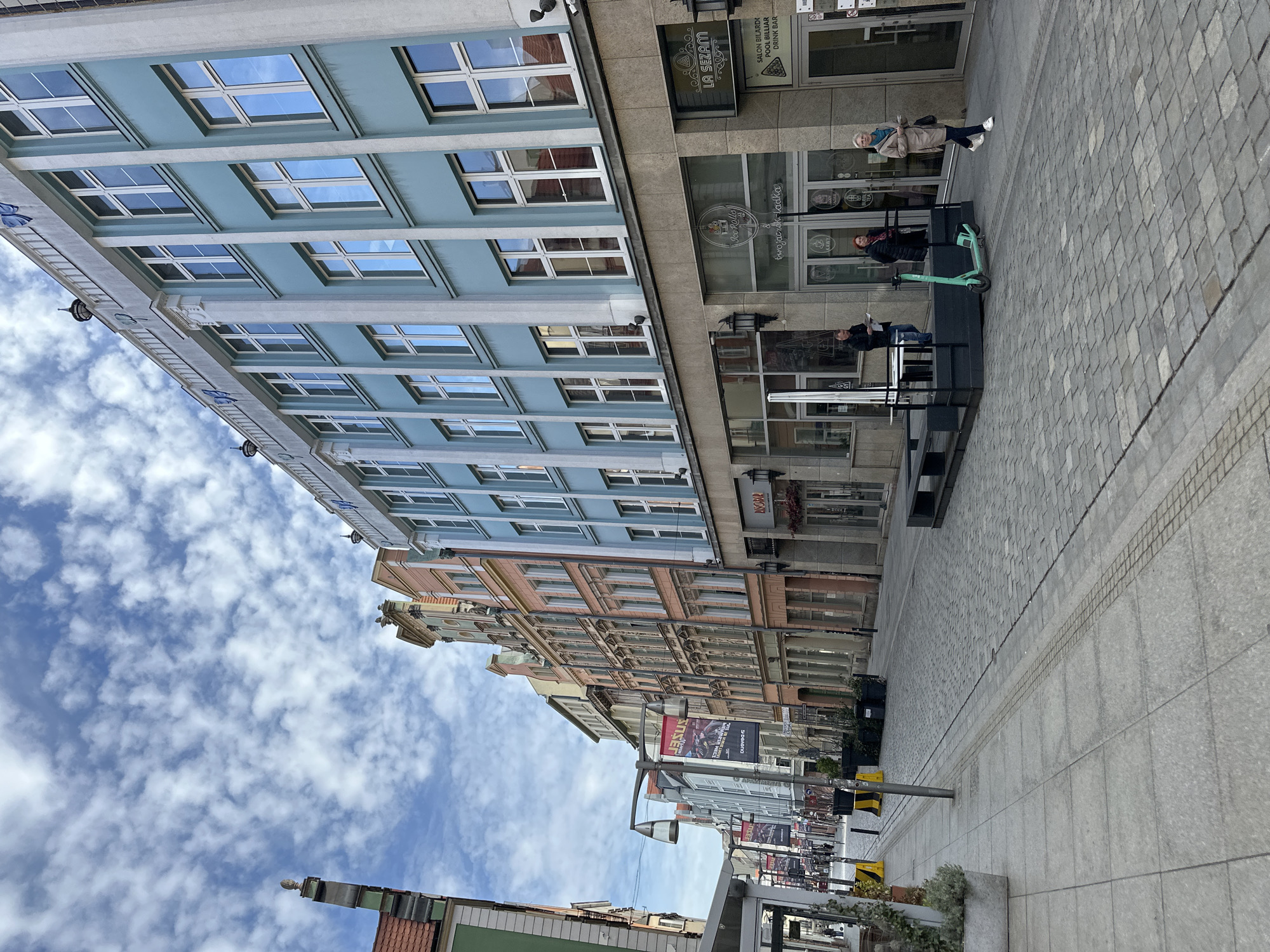 Here is a description of the image:
The photograph shows a street scene in Wrocław, Poland, angled upwards to capture the facades of the buildings along the street and a portion of the sky. The buildings are characterized by a mix of architectural styles, including a modern blue-colored building with many windows and older buildings with more ornate facades in pastel colors. The street is paved with stone, and there are some people visible, including individuals standing and one person on a green electric scooter. There are some store fronts along the street, including "LA SEZAM" and "LINOBAR". The sky is filled with white, fluffy clouds. There are street lights and bollards present along the sidewalk.