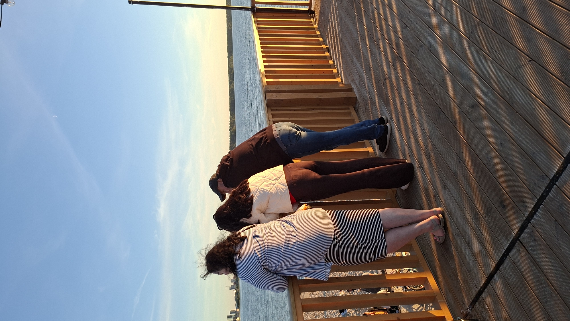 Three individuals are standing on a wooden pier, looking out at a body of water under a clear sky. The person on the left is a woman with dark curly hair wearing a striped long-sleeved shirt and a striped skirt. The person in the middle is a woman with dark hair wearing a quilted off-white jacket over a red shirt and dark trousers. The person on the right is a man wearing a dark hoodie and a baseball cap, with blue jeans and black shoes. The pier has wooden railings and the planks are illuminated by the low angle of the sun, casting long shadows. In the background, the water stretches to the horizon, where a tree line is visible. The skyline of Tallinn, Estonia, with several buildings, can be seen in the far distance. A crescent moon is visible in the sky. The overall scene suggests a late afternoon or early evening setting.