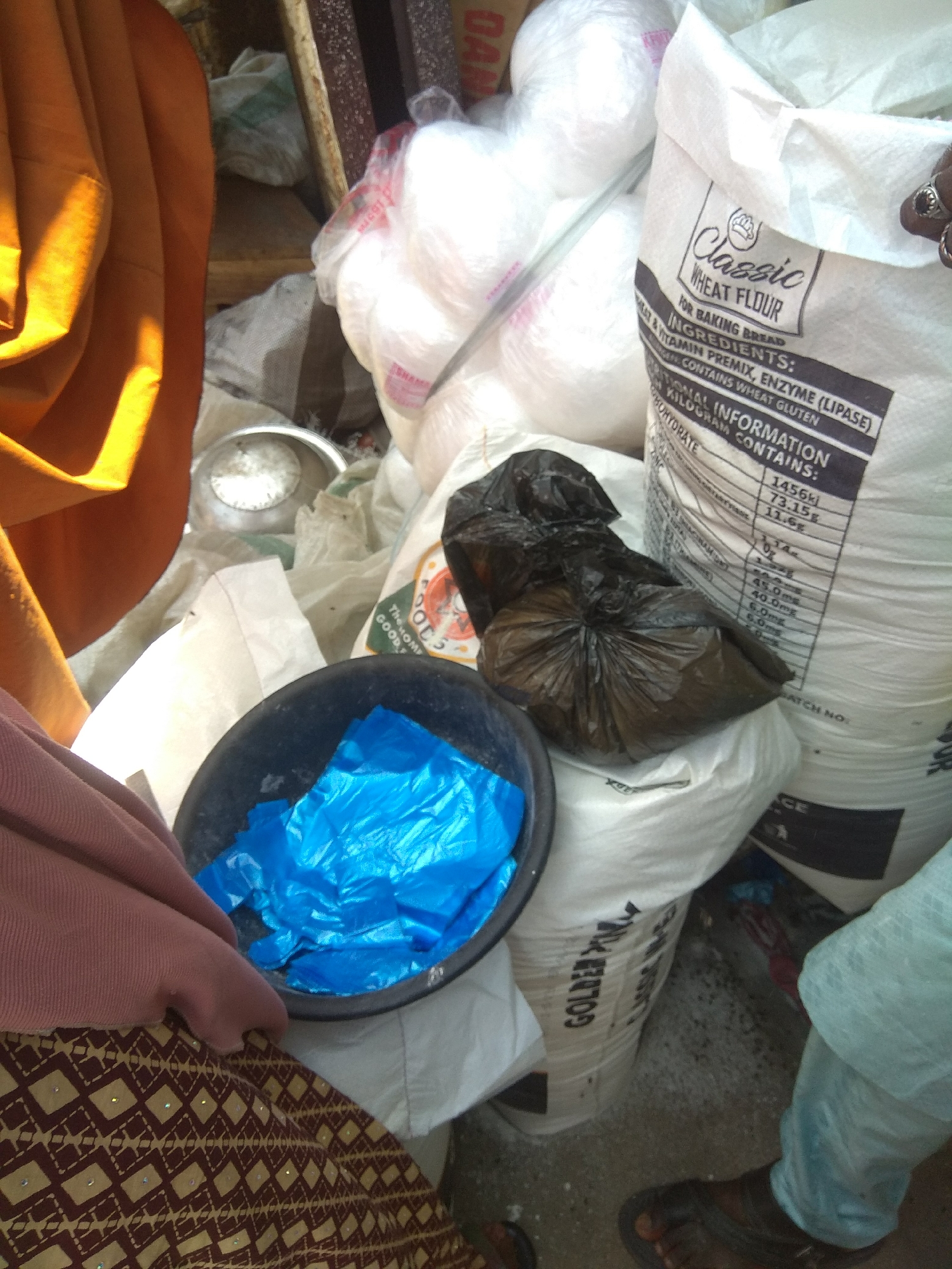 A medium shot captures a market stall scene in Jalingo, Nigeria, with a focus on sacks of flour and various food items. In the foreground, a dark bowl holds bright blue plastic bags, suggesting a distribution or packaging area. To the left, a person's legs are visible, clad in a pinkish-brown garment and a patterned brown and yellow cloth. Behind the bowl, a large white sack prominently displays "Classic Wheat Flour" and lists ingredients and nutritional information. Other sacks, some appearing to contain white granular substances, are stacked and arranged around the stall. The overall environment appears somewhat cluttered, typical of a busy market setting. The lighting is uneven, with strong sunlight hitting the left side of the image, creating bright highlights and shadows.