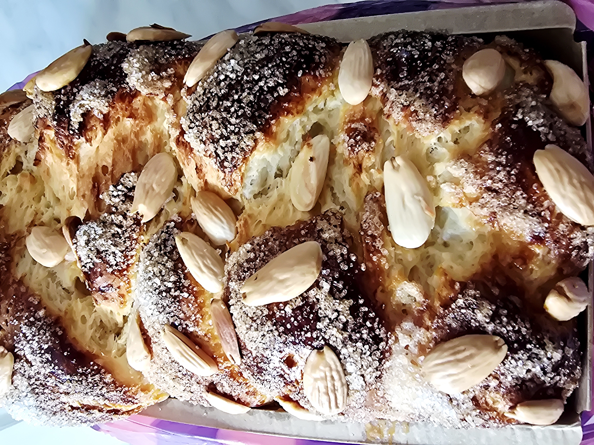 A close-up, eye-level shot captures a baked sweet bread, appearing to be Kozunak, a traditional Bulgarian sweet bread, richly adorned with sugar crystals and blanched almonds. The bread exhibits a golden-brown crust, indicative of a well-baked surface, interspersed with darker, caramelized patches. The sugar crystals provide a sparkling, textured coating over the crust, while the almonds are scattered irregularly across the surface, their pale color contrasting with the bread’s darker hues.
Given the appearance and the association of Kozunak with Bulgaria, this image might realistically be taken in Varna, Bulgaria, but there are no explicit visual cues to confirm the location.