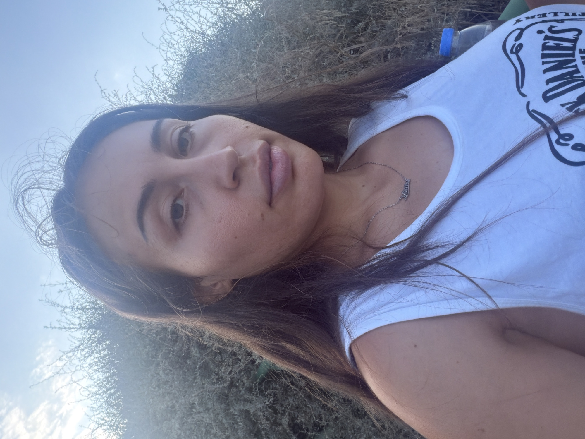 In Meneou, Cyprus, a young woman with long, dark brown hair is captured in a close-up selfie outdoors. She is wearing a white tank top, and a silver necklace with the word "Mama" is visible. Behind her, dry, shrub-like vegetation forms a dense background against a bright sky. A partially visible blue-capped water bottle is positioned to her right. The lighting suggests it is daytime.