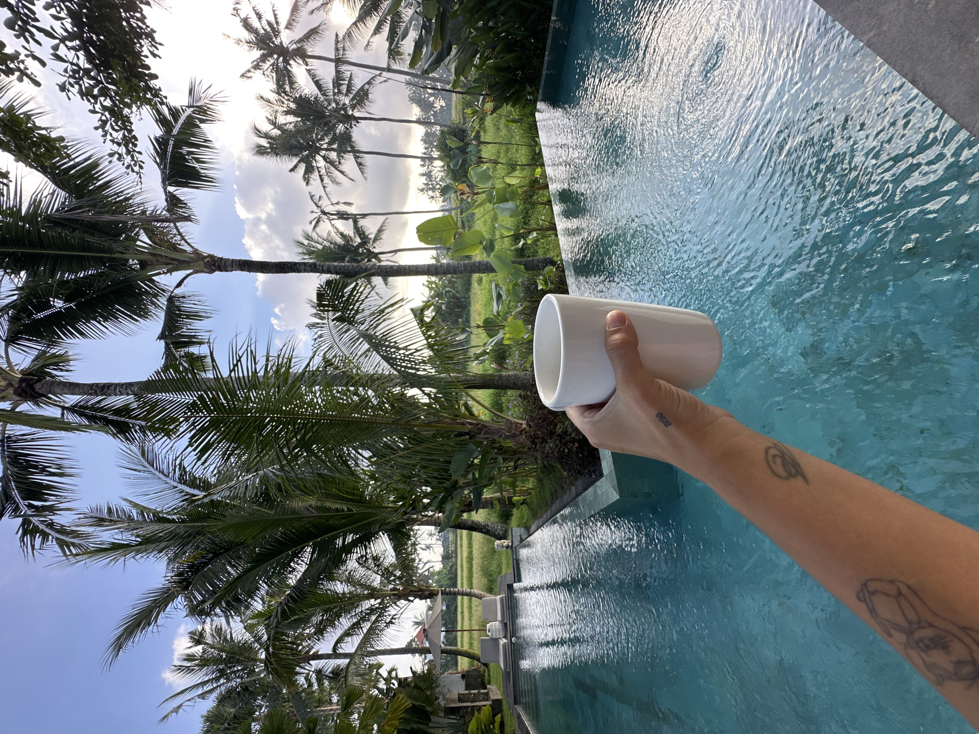 The image depicts a scene near a pool in Sukawati, Indonesia. The pool's turquoise water fills a portion of the frame. A person's arm, adorned with tattoos, extends into the frame, holding a white cylindrical cup. Palm trees line the background, framing a view of rice fields. The sky is partly cloudy with a mix of blue and white.