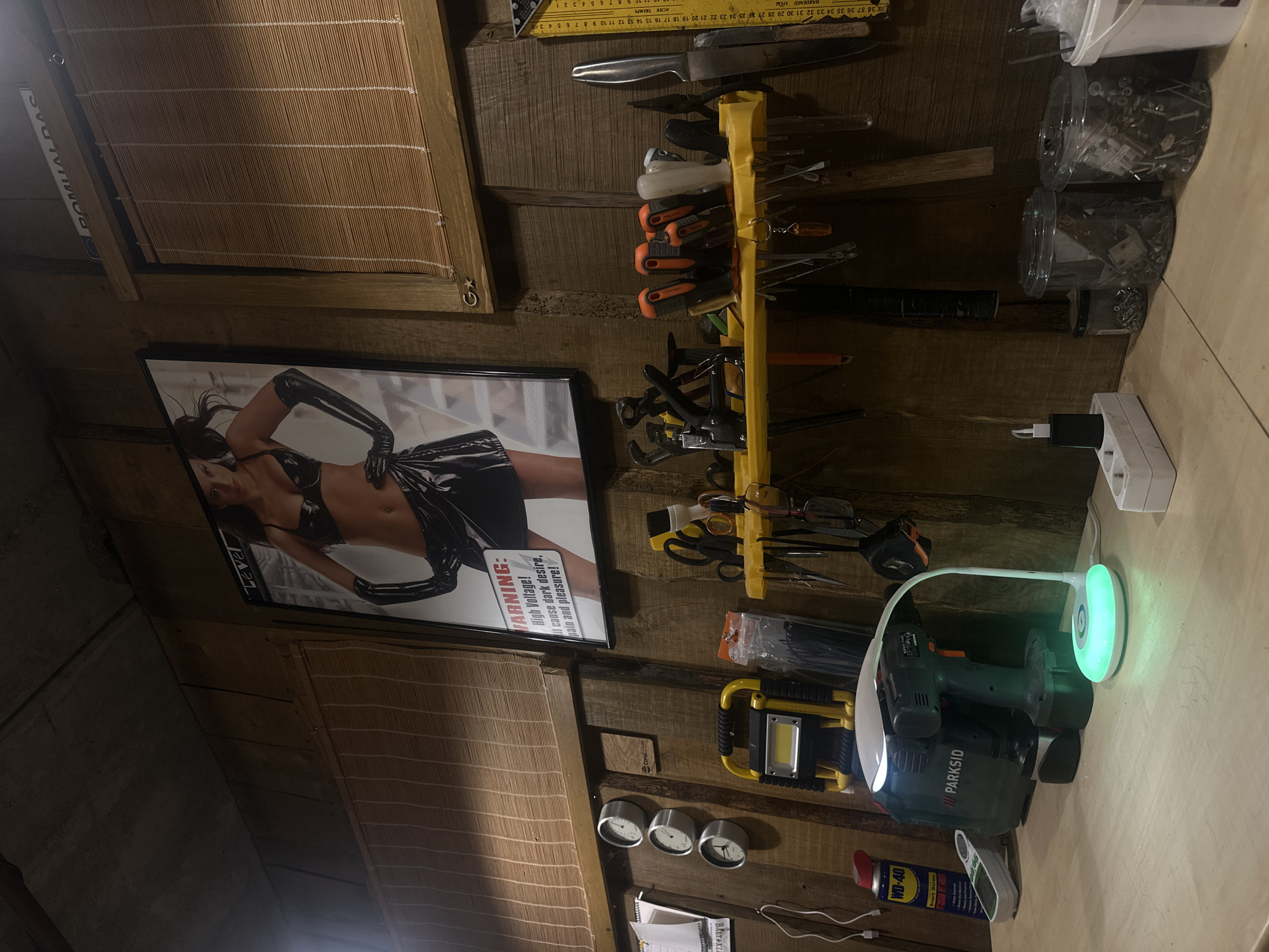 This is a candid, eye-level, medium shot of a workshop or garage space, illuminated by artificial light, with the camera angled slightly upwards and to the right. The room appears to be constructed from rustic wooden planks, giving it a distinctly workshop-like atmosphere.
Dominating the left side of the frame is a framed poster of a person wearing a black latex outfit, with text that reads "WARNING: High Voltage! I cause dark desire, pain and pleasure!". To the left of the poster, a slatted wooden blind hangs against the wall. The right side of the image showcases a collection of tools and equipment. A yellow tool organizer holds various items, including clamps, pliers, and scissors. Above this organizer, a metal ruler is attached to the wooden wall. Below the tool organizer, a yellow work light sits on a shelf.
Further to the right, on a wooden workbench, sits a power tool, possibly a drill, with a green and black casing that has the brand name "PARKSID" clearly visible. Next to it, a white desk lamp with a circular LED light source glows with a green hue. A white electrical outlet strip is plugged into the wall, with a black adapter inserted.
The overall impression is one of a functional workspace, possibly a garage or a dedicated DIY area. There are no people visible in the image, but the presence of tools, a poster, and various containers suggests it's a space used for work or hobbies. The lighting appears to be artificial, and the time of day is indeterminate. Several lines of text are visible, including the brand names "PARKSID" and "WD-40", the warning on the poster, and markings on the ruler. The context indicates the location is Lapės, Lithuania.