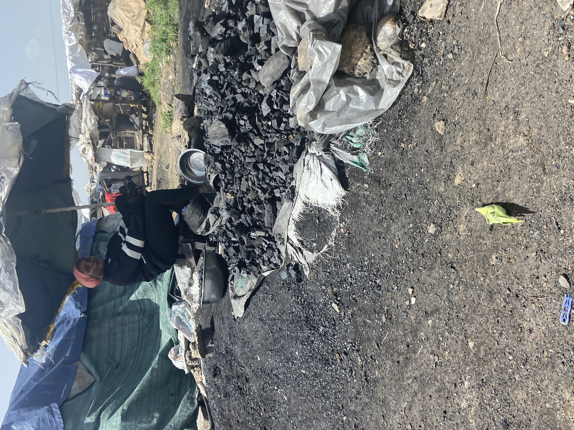 The image depicts an outdoor scene in what appears to be a marketplace or informal settlement in Bukuru, Nigeria. A person, wearing a pink headscarf and a black tracksuit with white stripes, is seated on a blue tarp. The person is looking up and to the left. Surrounding the person are various objects, including a large pile of charcoal, some of which is bagged in grey plastic. There are also tarps and what looks like makeshift structures in the background, suggesting a busy, possibly commercial, environment. A metal bowl is visible near the charcoal. The ground is a mix of dirt and charcoal dust. In the distance, more structures and some greenery can be seen. The lighting suggests it is daytime, possibly late morning or early afternoon, with bright sunlight casting shadows. A bright yellow object and a blue clothes peg are visible on the ground in the right portion of the image. The overall impression is one of a working environment, possibly a charcoal vendor's stall. There is no visible text in the image.