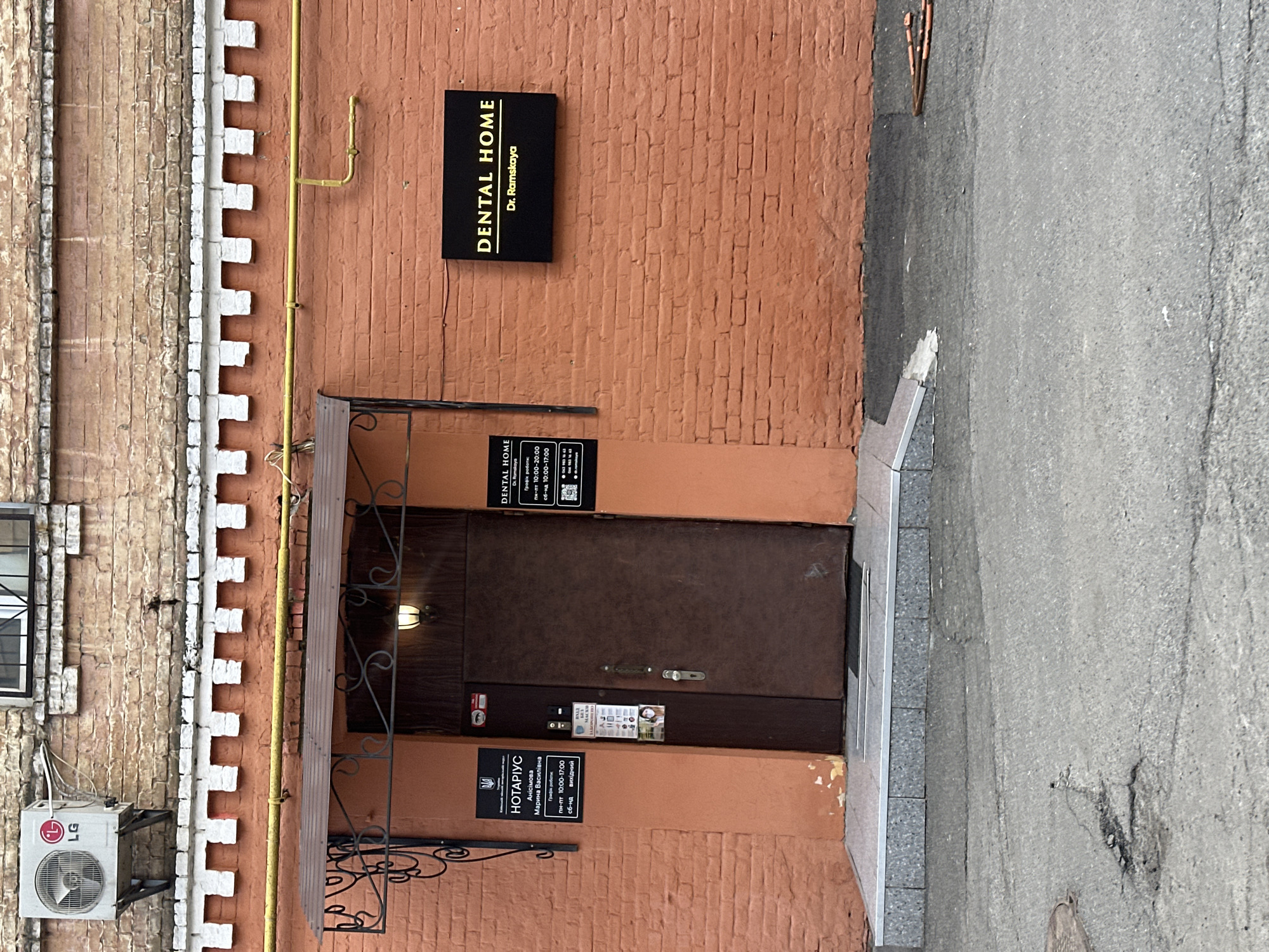The image displays the exterior facade of a two-story brick building in Kyiv, Ukraine. The lower portion of the facade is constructed of orange-red bricks, while the upper section, separated by a white crenellated molding, features lighter, weathered bricks. A central, dark brown metal door with a small, curved metal awning serves as the primary entrance. Above the door, beneath the awning, a single light fixture is visible.
To the left of the door, a black sign with the Ukrainian trident emblem identifies a notary service: "НОТАРІУС Анісімова Марина Василівна," with listed operating hours as Mon-Fri 10:00-17:00 and Sat-Sun closed. A smaller, vertical sign affixed to the door reads "ВХІД" (ENTRANCE) and "МАСКИ ЗАХИЩАЮТЬ" (MASKS PROTECT), accompanied by COVID-19 safety pictograms. To the right of the door, two identical black illuminated signs are mounted on the orange-red brick wall; both read "DENTAL HOME Dr. Ramskaya." The upper Dental Home sign details operating hours as Mon-Fri 10:00-20:00 and Sat-Sun 10:00-17:00.
An LG brand air conditioning unit is mounted on the upper, lighter brick facade on the far left. A yellow gas pipe runs horizontally across the orange-red brick wall, above the door awning, then angles downwards to the right. The entrance features a grey tiled threshold that transitions to a cracked asphalt pavement. A circular manhole cover is visible in the asphalt on the left side of the foreground. Several reddish-brown cylindrical fragments of debris are scattered on the asphalt near the building's base on the right. No individuals are present in the scene.