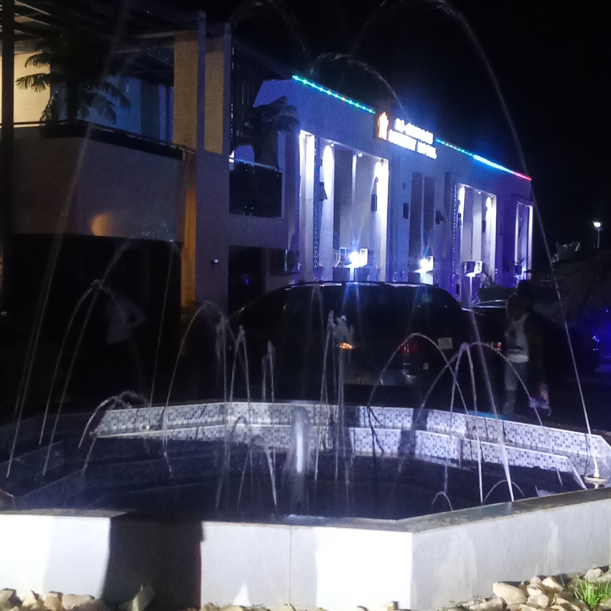 The image depicts a night scene in Akwanga, Nigeria, featuring a fountain in the foreground and a building labeled "M.A JOHNSON LUXURY HOTEL" in the background. The fountain is multi-tiered with a mosaic tile pattern. Water is spraying from multiple points around the fountain's edges. Behind the fountain, a dark vehicle is partially visible. People are indistinctly visible near the vehicle. The hotel is illuminated with white light highlighting the building's architecture, and a string of colored lights runs along the top edge. Palm trees are partially visible flanking the building. The ground around the fountain is covered in small rocks.