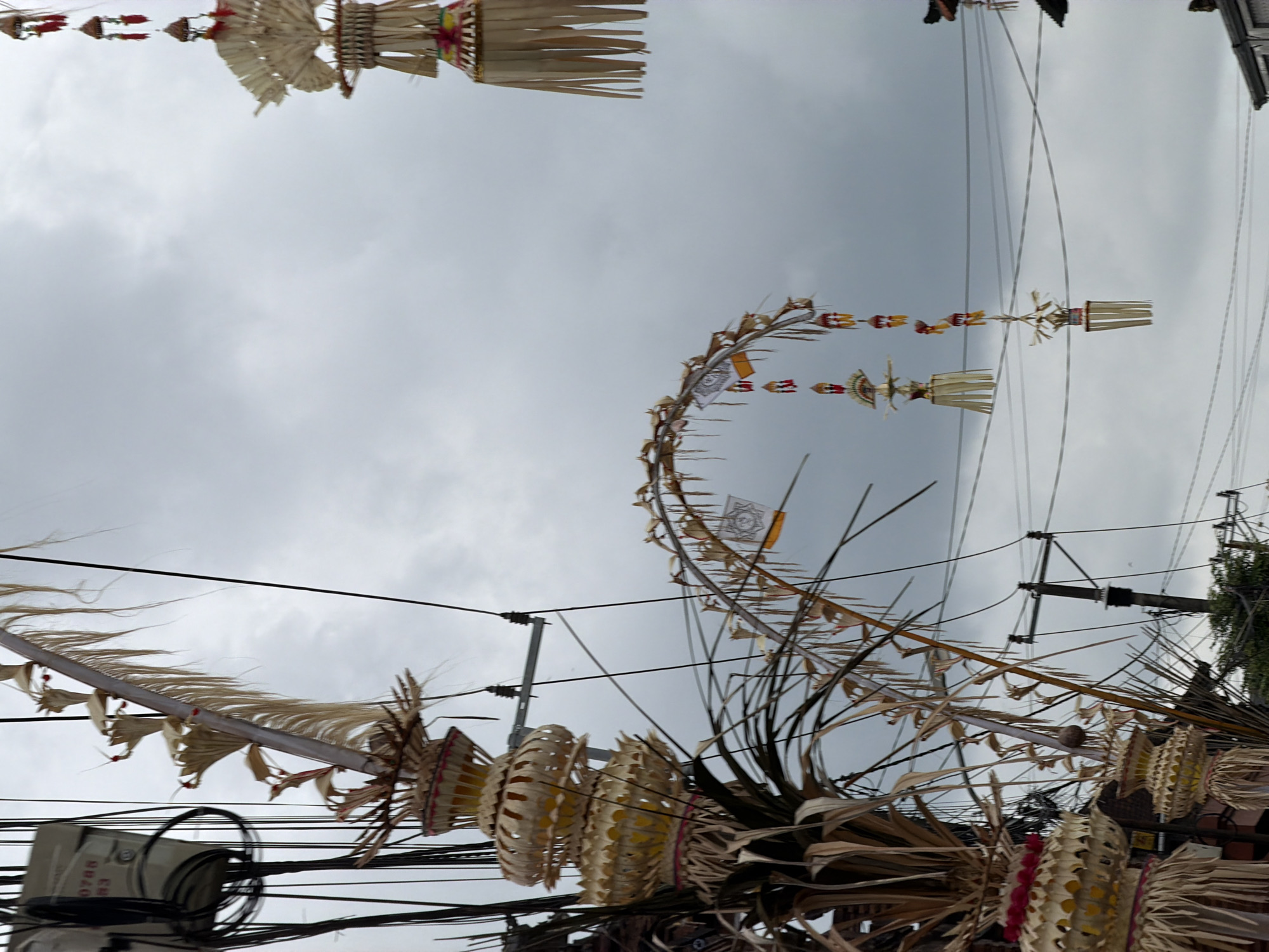 The image depicts an overcast sky above the city of Mengwi, Indonesia, with a focus on decorative religious offerings suspended from electrical wires. A prominent, multi-tiered decorative offering made of woven palm leaves and fabric is visible on the left side, cascading downwards. To the right, another similar, elaborate offering hangs from above. In the mid-ground, a curved arrangement of dried palm leaves adorned with smaller decorative elements and hanging offerings stretches across the frame, also suspended from wires. Several electrical poles and a network of wires are visible throughout the scene, indicating an urban environment. The overall impression is one of cultural or religious observance in a populated area, with traditional decorations integrated into the modern infrastructure.