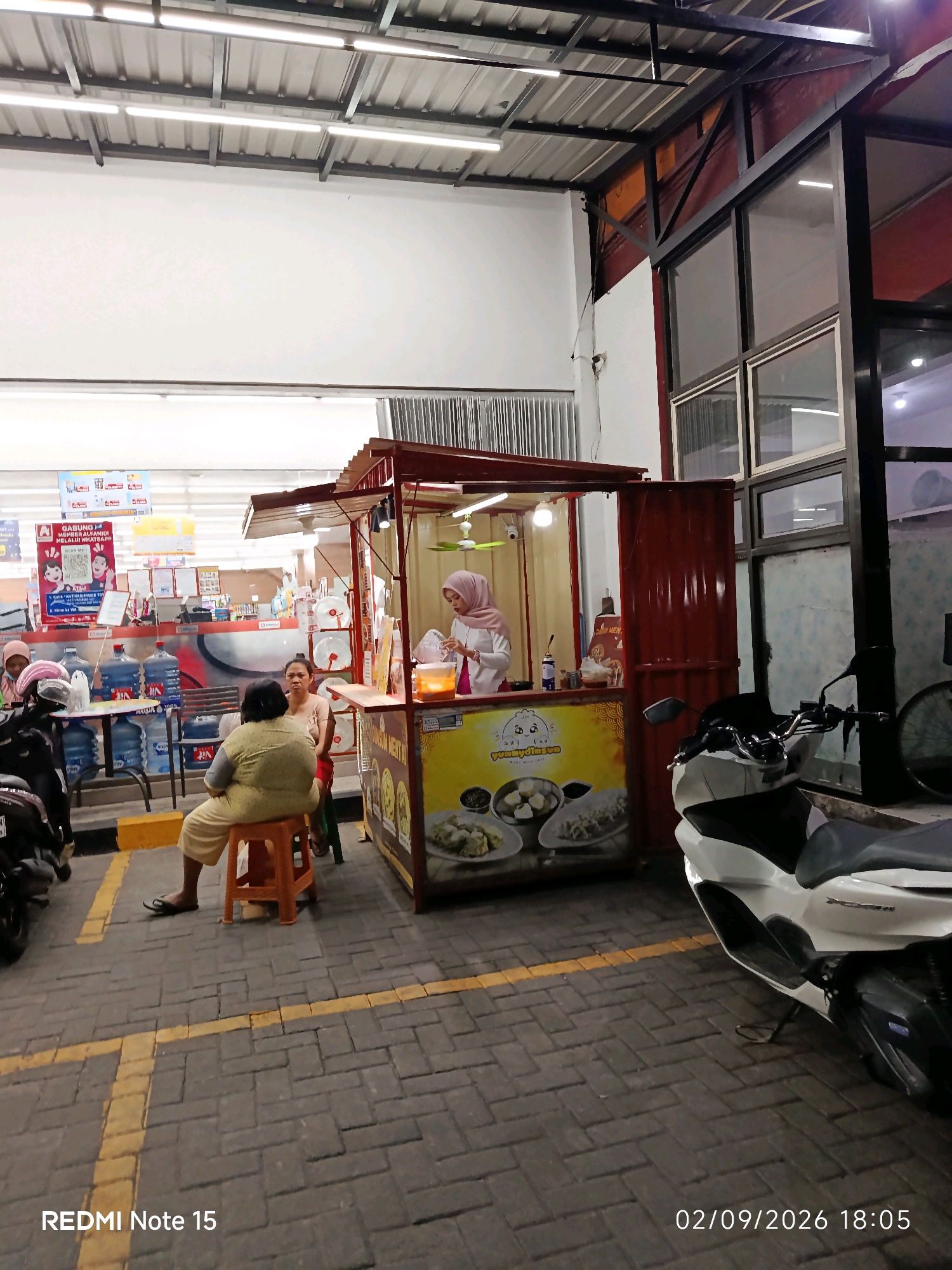 In Keboansikep, Indonesia, an outdoor food stall is set up in front of a store, as indicated by the "REDMI Note 15" watermark and the date "02/09/2026 18:05" in the lower right corner. The stall, made of red corrugated metal and a striped awning, displays images of food items like dim sum. A woman in a pink headscarf and white top operates the stall, serving customers. Two women are seated nearby; one in a yellow outfit sits on an orange stool, facing the stall, while another woman in a light brown top sits next to her, looking towards the stall. In the foreground, a white scooter is parked. The area is paved with interlocking stones, and yellow lines mark parking spaces. Water dispenser bottles are visible behind the seating area, and the store behind the stall appears to be a convenience store with brightly lit aisles.