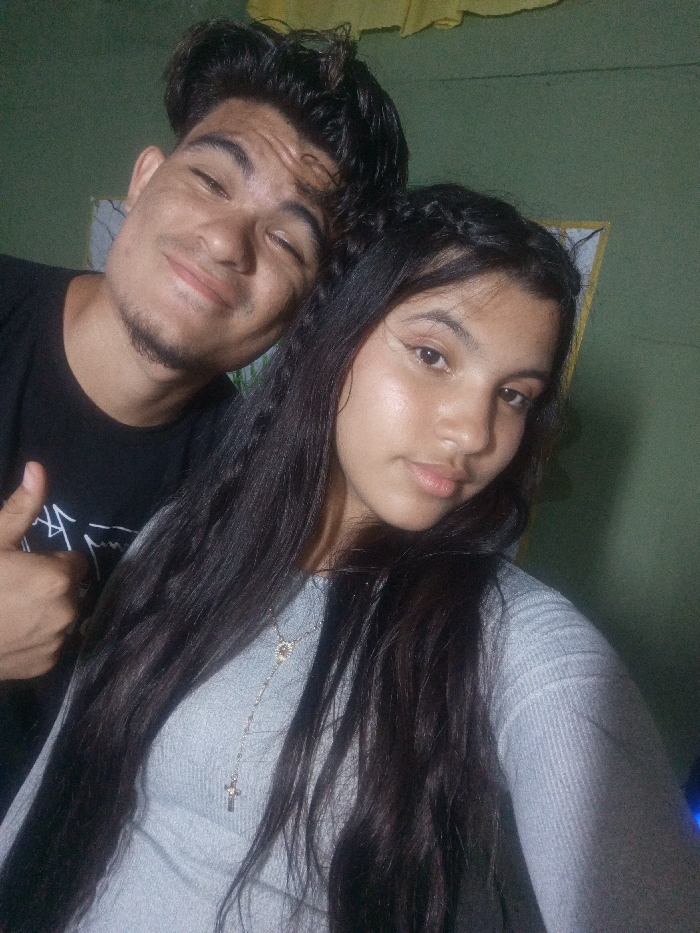 Two individuals are captured in a close-up indoor photograph taken in the city of Juan Griego, Venezuela. The subject on the left is a male, appearing to be a young adult, with light brown skin and dark hair styled upwards, exhibiting minimal facial stubble. He wears a black short-sleeved t-shirt, has his right eye partially closed or winking, and displays a slight smile. His right hand is visible in the lower left, making a "thumbs up" gesture. The subject on the right is a female, also appearing to be a young adult, with light brown skin and long dark hair, which includes some braids near the top and flows over her shoulders and chest. She is wearing a light gray ribbed long-sleeved top, adorned with a thin silver-colored necklace featuring a small circular pendant and a cross pendant. Her expression is neutral. The female is positioned slightly in front of the male. The background consists of a plain, dull green wall, with a partial view of a white-bordered rectangular object or frame and a piece of yellow fabric or paper visible in the upper right. The lighting indicates an indoor setting.