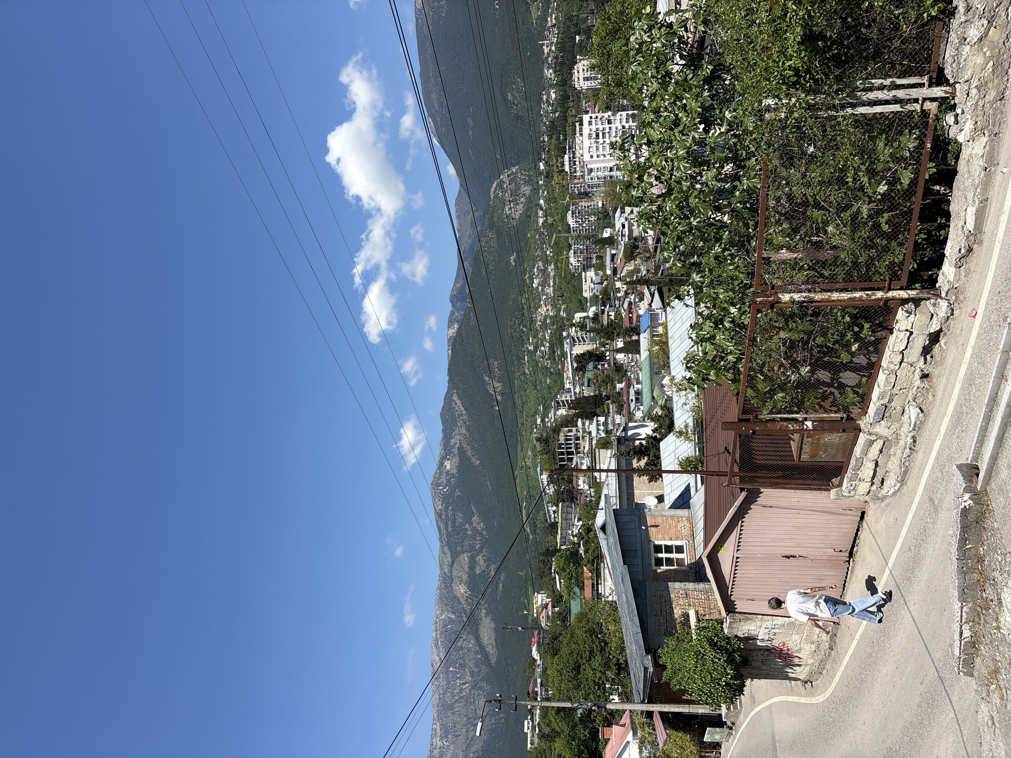 The image depicts a street scene in the city of Ялта, Ukraine. A man wearing a white shirt and blue jeans is walking on a paved road, close to the buildings. The surrounding environment consists of buildings of varying heights and architectural styles, some residential, set against a backdrop of a mountain range. There are power lines visible in the sky, against a bright blue sky with some white clouds. Some metal fencing is visible along the roadside. Vegetation, including trees and bushes, are interspersed among the buildings and alongside the road.