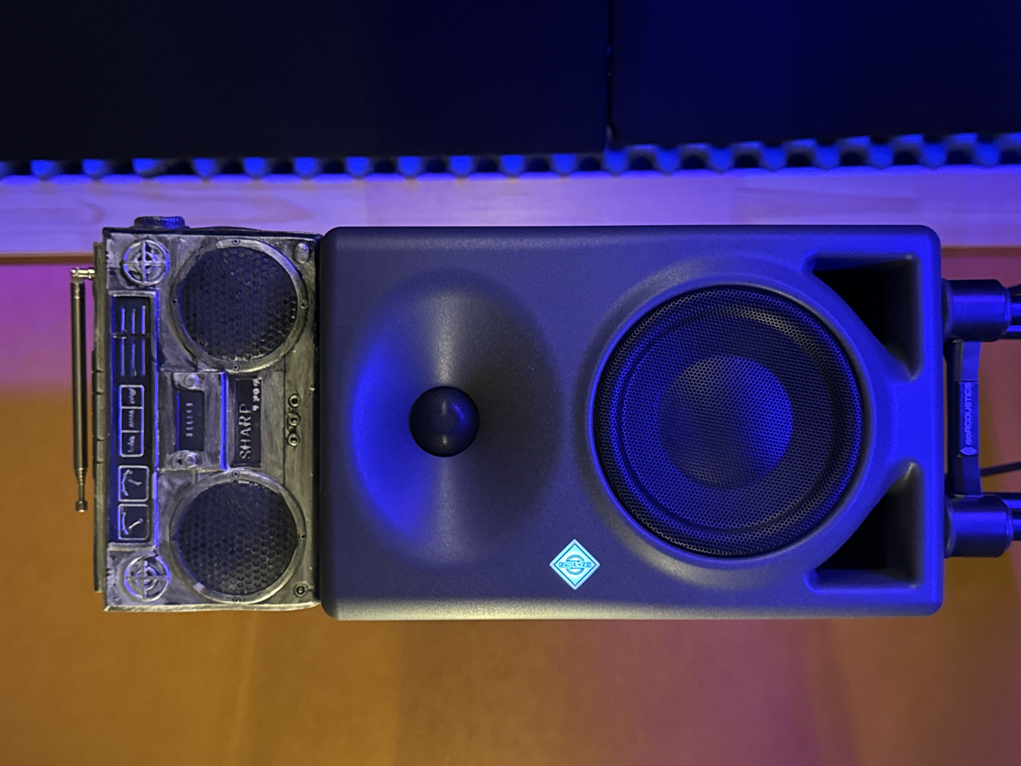 A top-down view showcases a sound system on an orange surface. To the left is a modified, steampunk-style Sharp boombox with dual speakers and an antenna. To the right is a gray Neumann studio monitor with a circular speaker and the Neumann logo. The speaker is mounted on a dark stand. The background features dark acoustic panels, lit with blue LED lights. There is no indication of the location being Plovdiv, Bulgaria within the image itself.