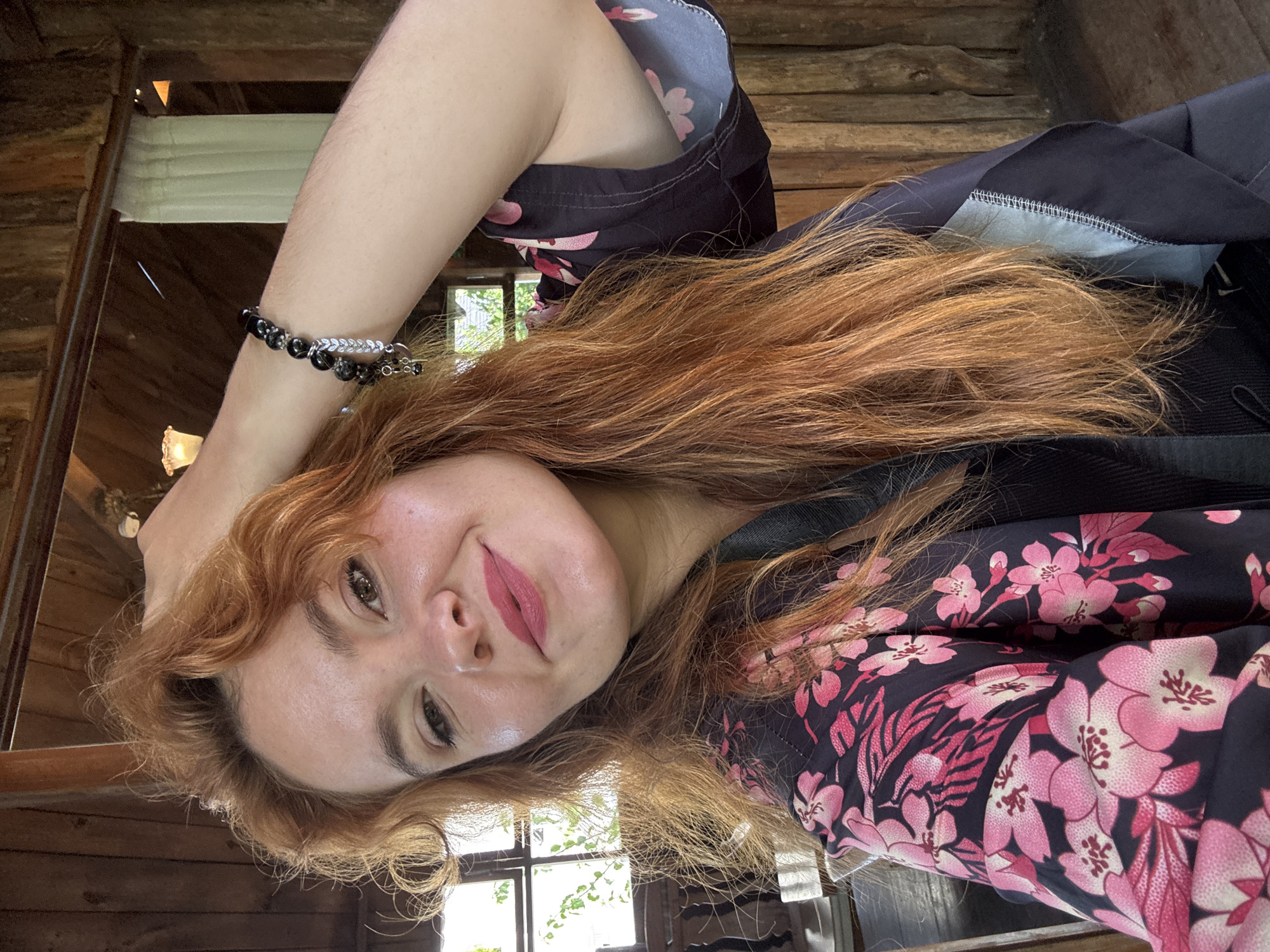 The image shows a woman with long, auburn hair, wearing a dark robe with a pink floral pattern. She is smiling and has her hand raised to her head. She is wearing a bracelet on her wrist. The background appears to be a rustic wooden interior, possibly a restaurant or cafe, with natural light filtering in from a window. The overall setting suggests a relaxed and casual atmosphere. The image does not provide specific information about the city of Da Lat, Vietnam.