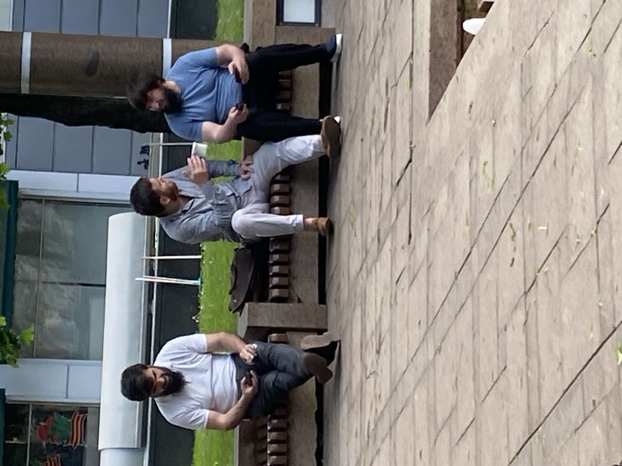 Three bearded men are sitting on a stone bench in an outdoor setting. One man wears a blue shirt and black pants, another wears a light gray button-down shirt and light gray pants, and the third wears a white shirt and dark gray pants. All three men are holding mobile phones. The bench is made of dark brown stone. The ground around the bench is paved with stone tiles. In the background are buildings and greenery. The location is likely Moscow, Russia, based on architectural styles and urban landscape elements.