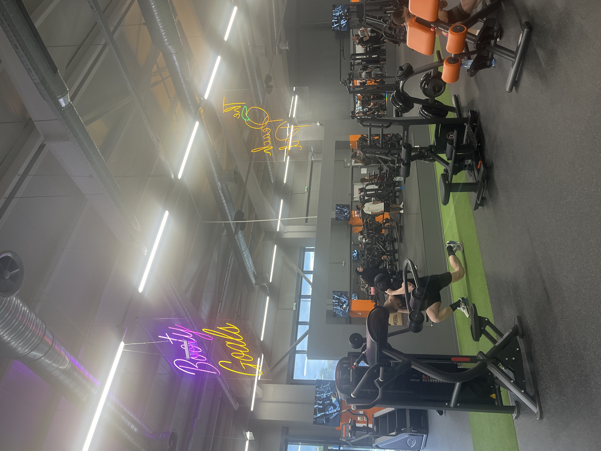The image depicts the interior of a gym, likely located in Plovdiv, Bulgaria. The gym features rows of exercise equipment, primarily weight machines, arranged along a gray floor with green sections. Several individuals are engaged in various workout activities. Fluorescent lights illuminate the space, and there are neon signs with phrases such as "Booty Goals." Overhead, ventilation ducts and support beams are visible.