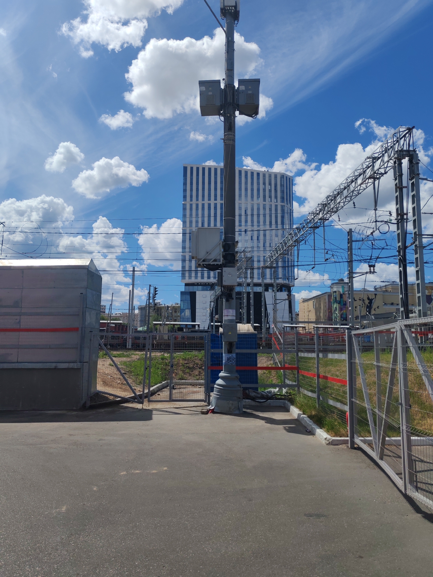 This is a photograph taken outdoors on a sunny day with a bright blue sky and scattered white clouds. The foreground is dominated by a paved surface, possibly a pathway or road, leading towards a series of metal fences and a large, metallic industrial-looking structure on the left.
Dominating the center of the image is a tall, dark gray pole with various equipment attached, including what appear to be lights and boxes. This pole is positioned in front of a modern, tall building with a facade of vertical blue and white stripes. Behind the fenced area and the central pole, there are railway tracks and overhead power lines, suggesting a railway station or a similar transportation hub. Further in the background, several other buildings can be seen, one of which is yellow and has colorful graffiti on its side.
The overall scene appears to be a blend of urban infrastructure and natural elements. The presence of the railway and the modern office building points to an urban environment. The clear sky and bright sunlight indicate it is likely daytime, possibly late morning or early afternoon. There are no people visible in the image. The mood is neutral, conveying a sense of everyday functionality and the contrast between man-made structures and the open sky. There is no visible text in the image.