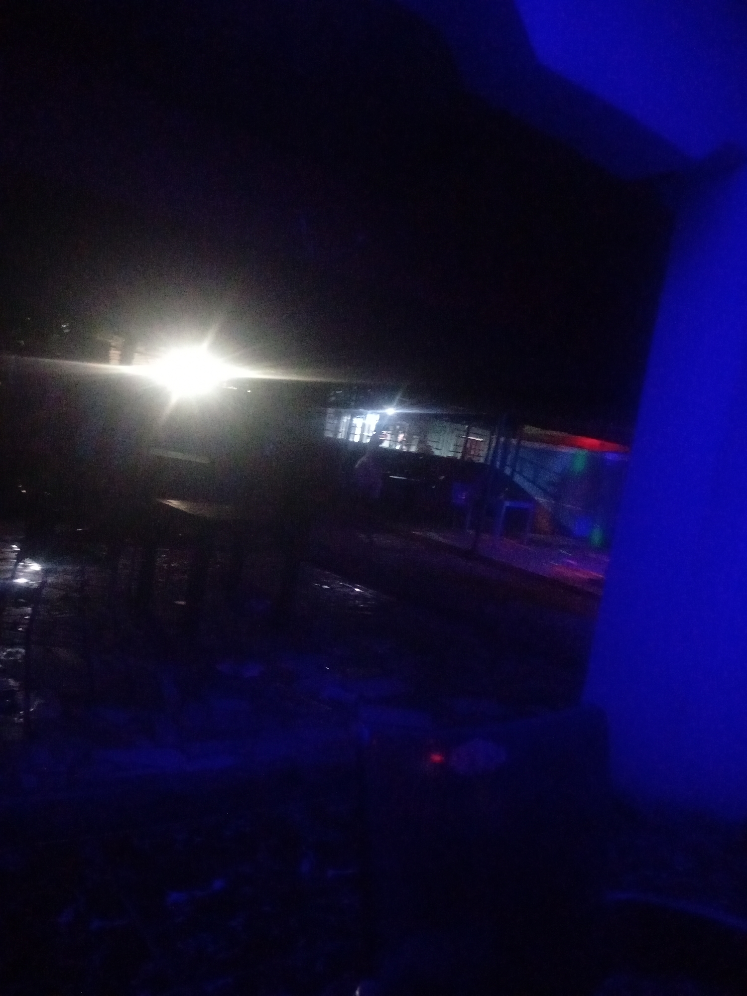 The image depicts a dimly lit outdoor scene at night. A bright light source, possibly headlights from a vehicle, illuminates the left side of the frame, casting long shadows. In the background, there appears to be a structure with windows, some of which are lit. The ground in the foreground is wet and reflective, suggesting recent rain. Blue lighting emanates from the right side of the image, creating an atmospheric effect. The overall impression is that of a public space, possibly a street or an open area near a building, in Sabon Gari, Nassarawa, Nigeria, at night.