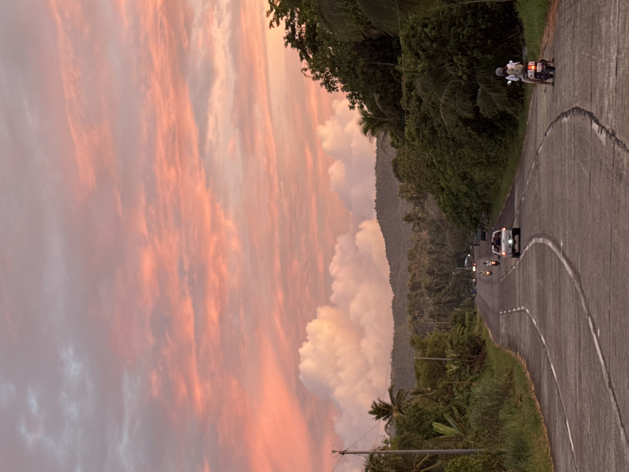 The image depicts a sunset view on a road in the city of Pilar, Philippines. The sky is filled with pink and orange clouds. A road curves through the image, with lush green vegetation on one side and a mountain range in the background. Vehicles, including a car and a motorcycle with two riders, are present on the road.