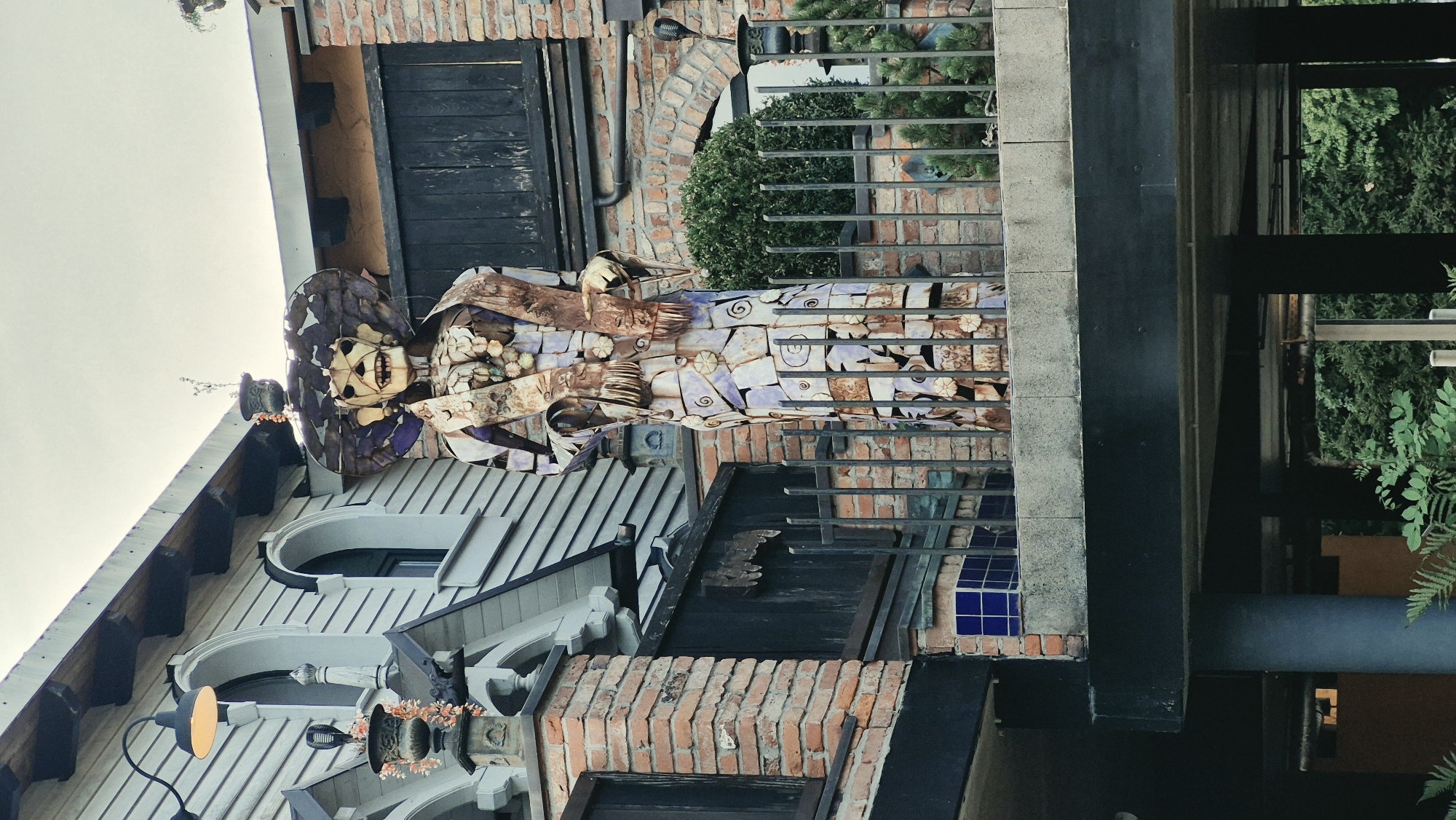 The image shows a large, stylized sculpture of a skeleton wearing a wide-brimmed hat and a long dress. The sculpture appears to be made of metal and possibly ceramic tiles, with a skeletal face and outstretched arms. It is standing on a balcony behind a metal railing. In the background, there is a building with brick and wooden elements, as well as arched windows and a hanging lamp. Lush green foliage is visible at the bottom of the frame. The sculpture's artistic style evokes themes of Day of the Dead or similar cultural celebrations. This sculpture is located at the entrance to the "Welten” garden at the Chemnitz Zoo in Chemnitz, Germany.