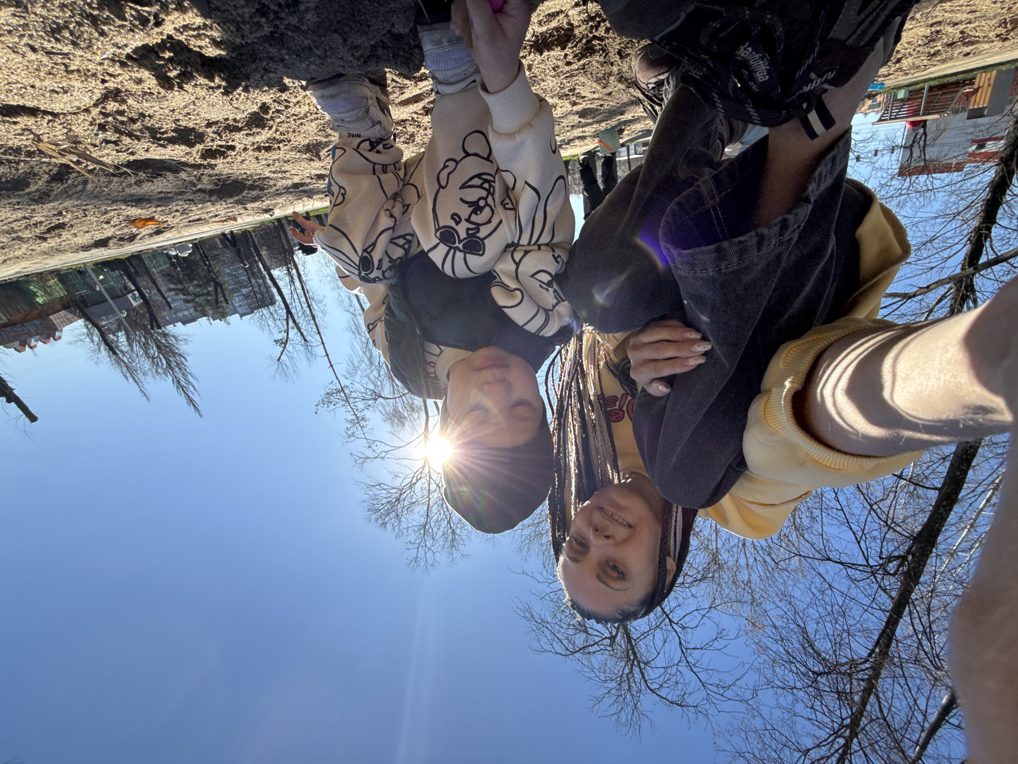 Two individuals are pictured in a selfie taken at a low angle, with a bright blue sky and bare tree branches in the background. The sun is visible behind the individual on the left, creating a starburst effect. The ground, which appears to be a dirt or sand surface, is in the background, and a few buildings are discernible in the far distance.
The individual on the left is wearing a dark-colored hat and a patterned beige and black sweatshirt. The person on the right is wearing a yellow sweatshirt under a dark-colored jacket, and their hair is styled in braids. The location appears to be a park or outdoor area in Zhukovo, Russia, as suggested by the architectural style of the buildings visible in the distance, as well as the type of trees present. The individuals are interacting by posing for the selfie.