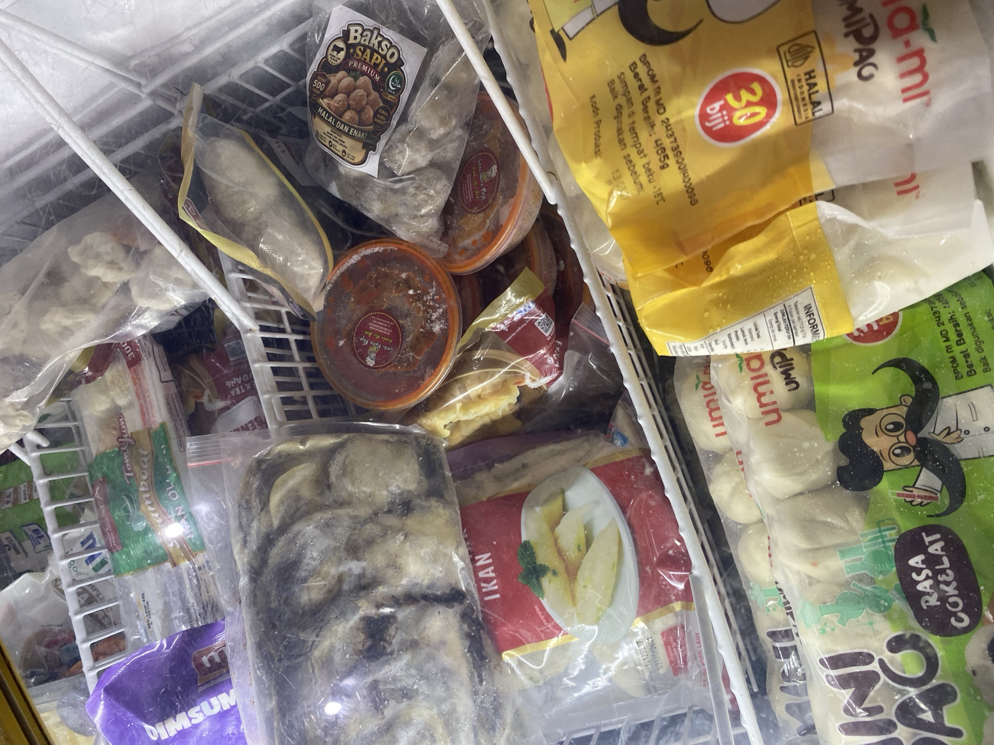The image is a close-up, overhead shot of a freezer display case filled with various frozen food products. The products are packaged in plastic bags and containers, with some labels visible in Indonesian. Among the visible items are bags of what appear to be fish, meatballs, and some form of dough or buns. Some products have cartoon illustrations on their packaging. The freezer shelves are made of white plastic-coated metal. The lighting suggests an indoor commercial setting. There are no people visible in the image. The overall environment appears to be a retail freezer in Indonesia.