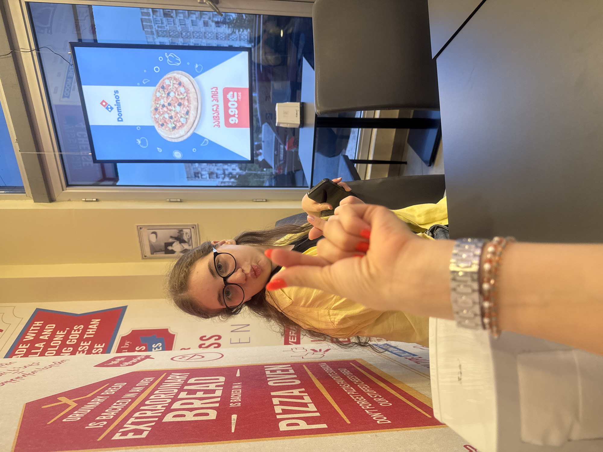 The image shows a young woman with glasses and long brown hair, wearing a yellow shirt. She is looking at the camera and making a "rock on" hand gesture with her right hand, while her left hand is partially visible in the foreground. She has bright orange nail polish on her fingernails.
The scene appears to be inside a Domino's Pizza restaurant in Tbilisi, Georgia. A large screen displaying a Domino's advertisement with a pizza and the price "9.90" is visible in the background through a window. The screen has Georgian text on it, including "პადანა პიცა".
There is also a large cardboard box with text on it, which appears to be related to pizza or bread. Some of the visible text on the box includes "ORDINARY BREAD", "EXTRAORDINARY BREAD", and "PIZZA OVEN".
The overall impression is that the woman is posing for a photo while at the restaurant, possibly waiting for or eating pizza. The lighting suggests it is daytime.