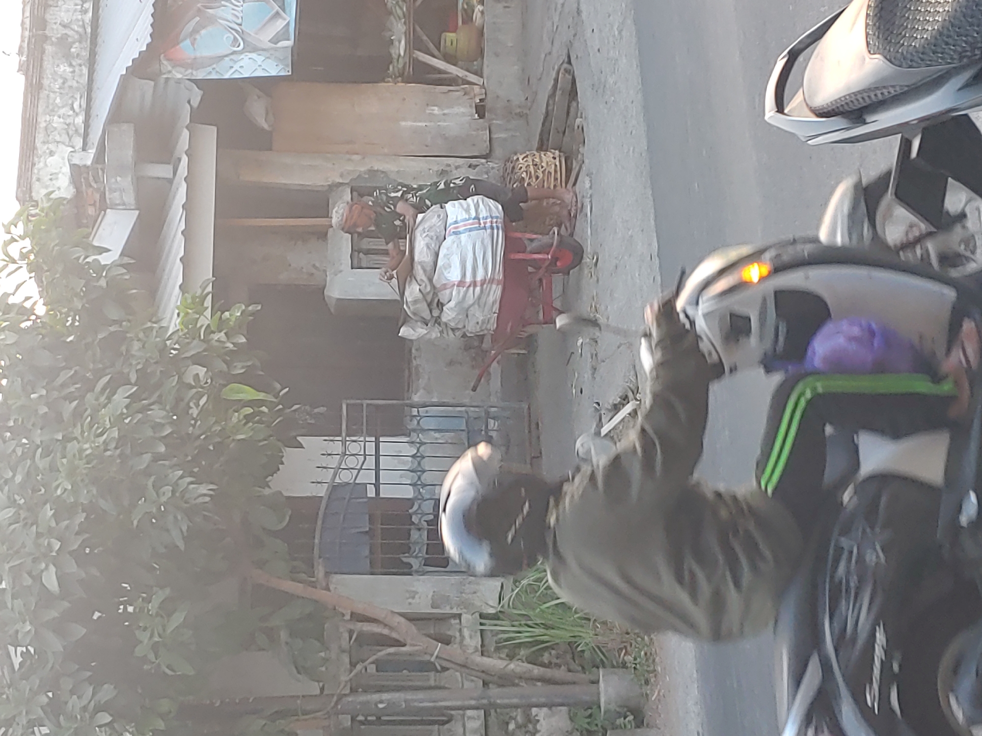 A medium shot captures a street scene in Kembang Kerang, Indonesia, during daylight. In the foreground, a person wearing a dark helmet and olive green jacket rides a black and silver scooter. They are wearing black pants with green stripes down the sides. The scooter has a purple bag hanging from the front. In the mid-ground, a person in a patterned shirt and dark pants is near a red wheelbarrow loaded with large sacks. They appear to be interacting with the sacks. Behind them is a building with a corrugated metal roof and a gate. A tree with green leaves is on the left side of the image. The ground is paved.