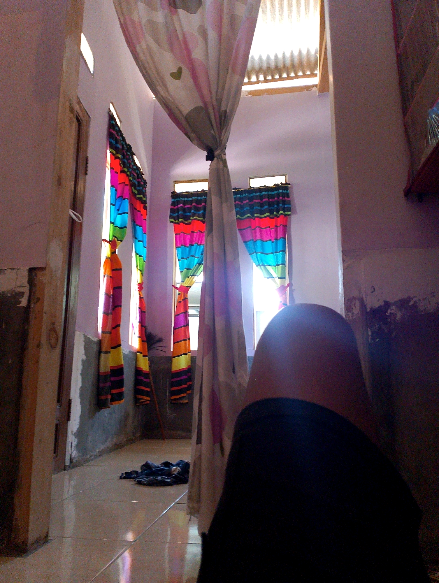 The image depicts an interior scene, likely a hallway or living space, in a building located in Desa Pagandaran, Indonesia. The room features light purple walls and a tiled floor that reflects light. Several windows are adorned with vibrant, multi-colored curtains, with vertical stripes in shades of pink, orange, yellow, blue, and green. One window also has a more subdued curtain with black, pink, blue, and green horizontal stripes. A large, light-colored curtain hangs in the foreground, partially obscuring the view. In the lower right quadrant, a dark, indistinct shape, possibly a person's leg covered by dark clothing, is prominent. On the floor, near the center left, a dark pile of fabric lies. The overall impression is of a lived-in space with colorful decor.