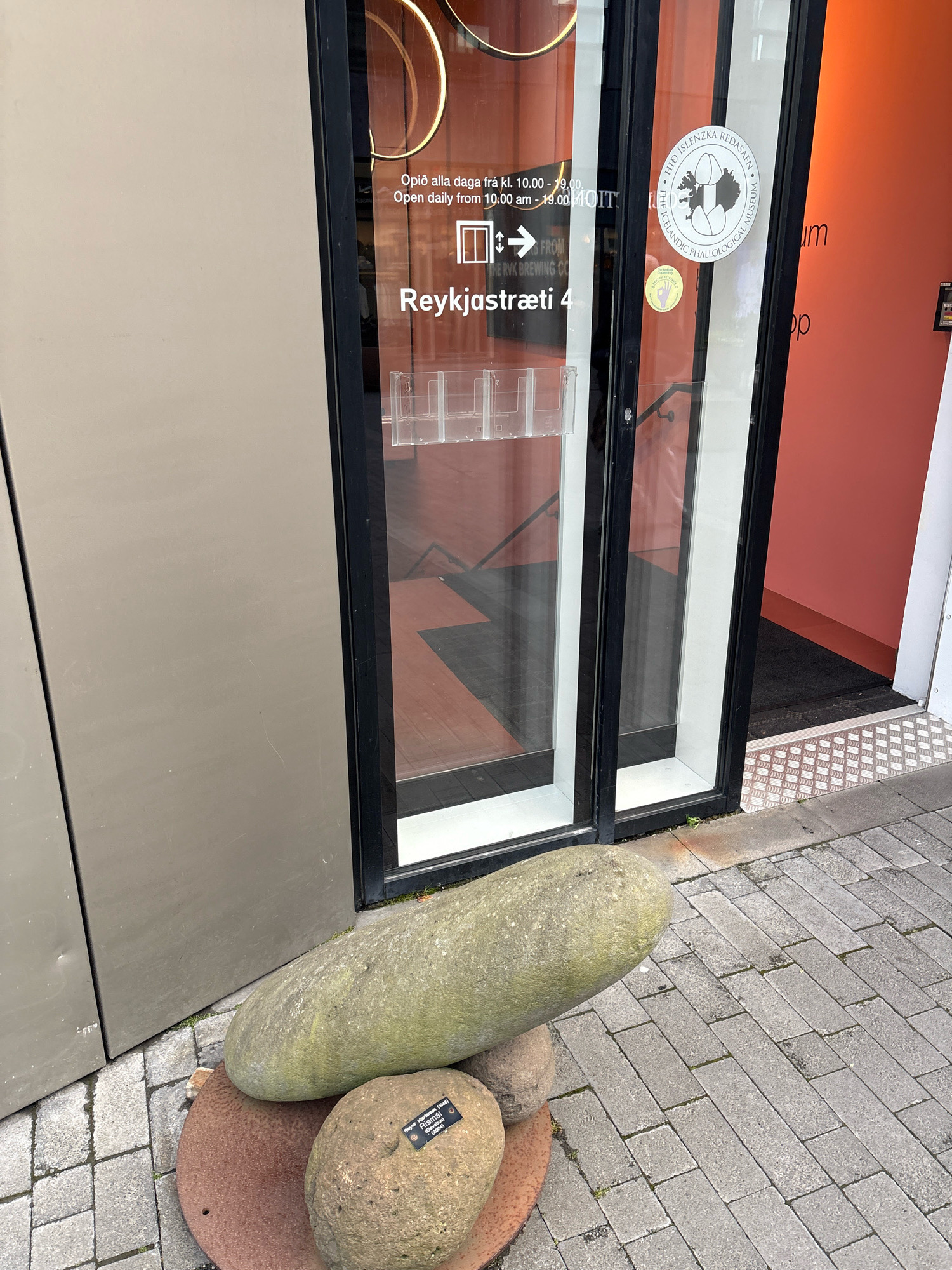 The image shows the entrance to the Icelandic Phallological Museum, also known as the "Phallological Museum" or "Pinnacle of Man", located at Laugavegur 116 in Reykjavik, Iceland. The museum is housed in a building with a large glass storefront, revealing the interior with displays and signage. The exterior features a prominent sign that reads "Reykjavikstræti 4," indicating the museum's address. Outside the entrance, large, smooth stones are displayed, serving as an artistic or decorative element. One of the stones has a small plaque attached to it. The ground is paved with cobblestones, and a metal ramp is visible near the entrance. The overall scene suggests a public space in a commercial area of Reykjavik.