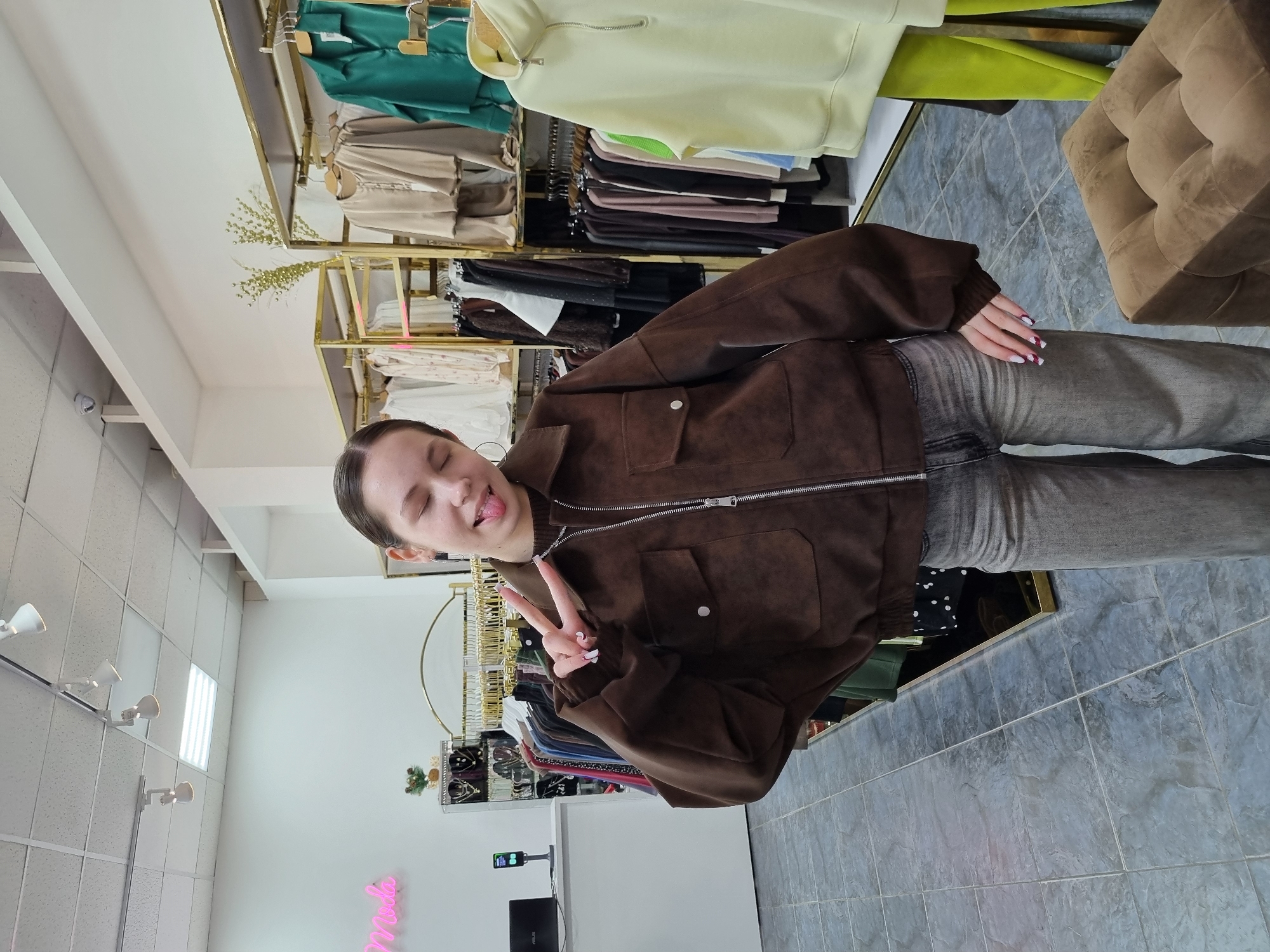 The image displays the interior of a clothing store situated in Chaikovsky, Russia. A young adult female stands centrally, facing the camera with her eyes closed and tongue slightly extended. She is wearing a brown, long-sleeved zippered jacket featuring chest pockets, a dark top underneath, and grey wide-leg jeans. Her right hand forms a peace sign, and her fingernails exhibit white and red tips. Hoop earrings are visible on her ears.
The environment includes white walls, a grey tiled floor, and a white ceiling equipped with track lighting and a rectangular LED panel. To the left, a white counter supports a black ASUS laptop and a black vertical device, likely a payment terminal or scanner. A pink neon sign partially spells "Moda" on the back wall. Gold-colored clothing racks are visible, laden with various garments including shirts, sweaters, and dresses in shades of beige, green, white, and brown. Displays containing jewelry, specifically necklaces, are positioned behind the counter. A brown tufted ottoman is located in the lower right foreground. A white security camera is mounted near the ceiling in the upper left background.