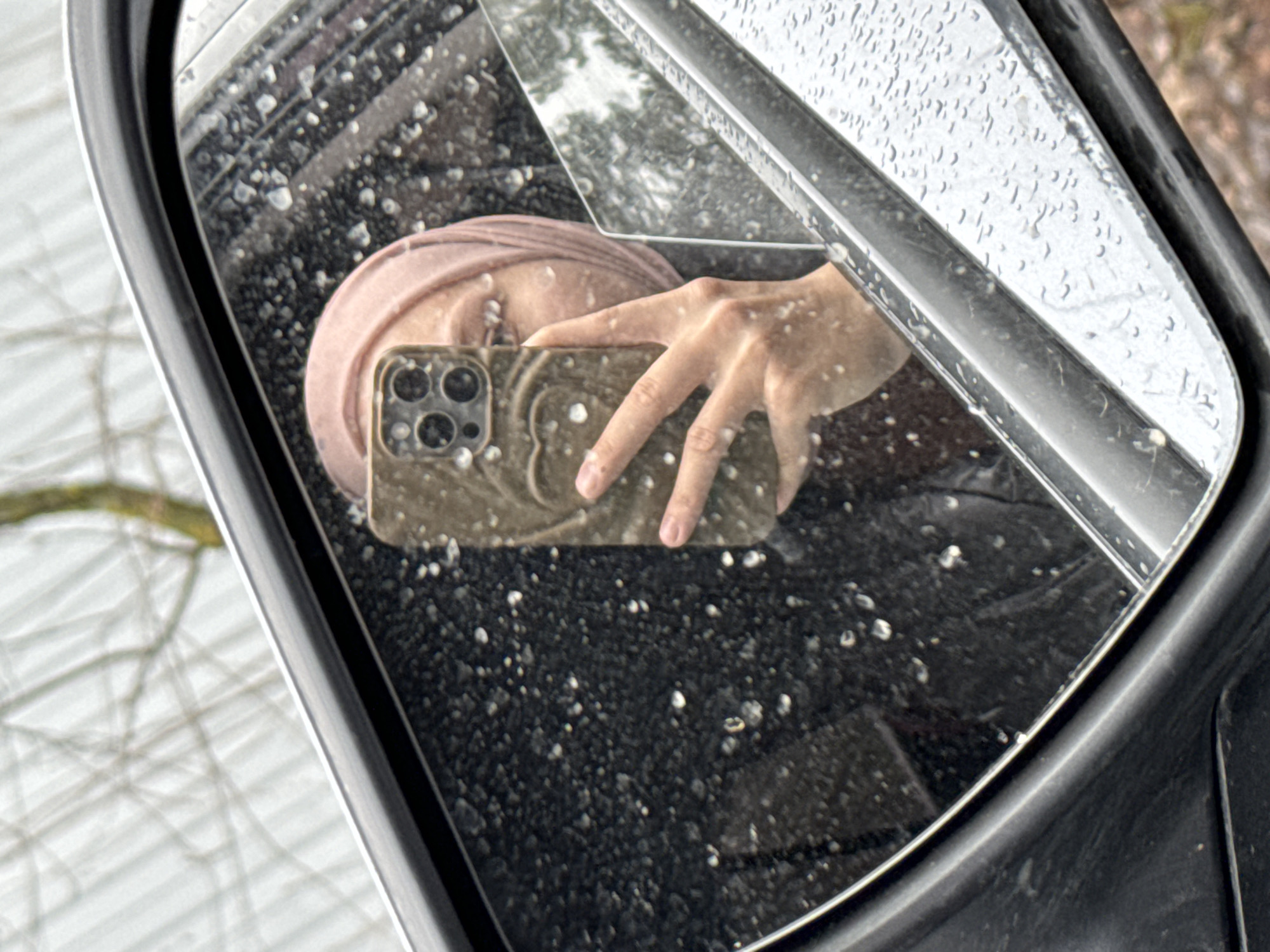 A reflection in a car's side mirror captures a person taking a selfie. The person wears a pink head covering and holds a smartphone with multiple camera lenses. The mirror surface is covered in water droplets. The background visible in the mirror is blurry, but an exterior scene can be discerned, possibly reflecting trees. Based on the presence of visible water droplets, the environmental condition is likely rainy. No specific landmarks are discernible to definitively pinpoint the location as Chaikovsky, Russia. More visual cues within the scene would be required to confirm the location.