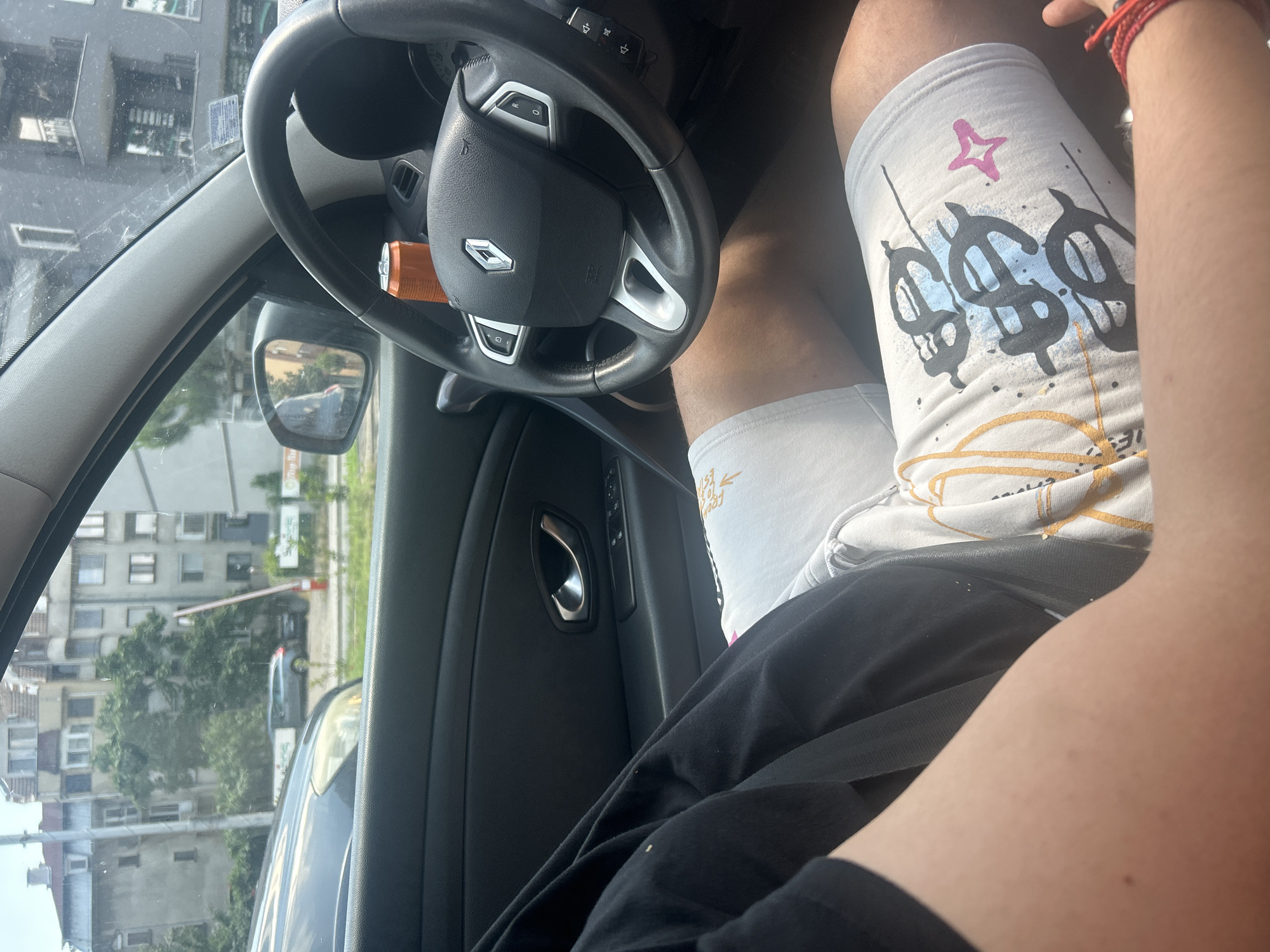 The image depicts the interior of a car, viewed from the perspective of a passenger. A person's legs are visible, wearing white shorts with a black and blue dollar sign graphic print and a small pink star. The person's right arm is extended and resting on their leg, with a red string bracelet on their wrist. In the foreground, on the car's dashboard, is an orange soda can. The steering wheel, which has a Renault logo, is visible in the right side of the frame, along with various car controls. Outside the car's window, a street scene can be observed, with buildings and trees. The presence of buildings with a distinct architectural style and the overall urban setting are consistent with the city of Plovdiv, Bulgaria. The environment appears to be a street or parking area within the city. The time of day seems to be daytime, given the natural lighting.