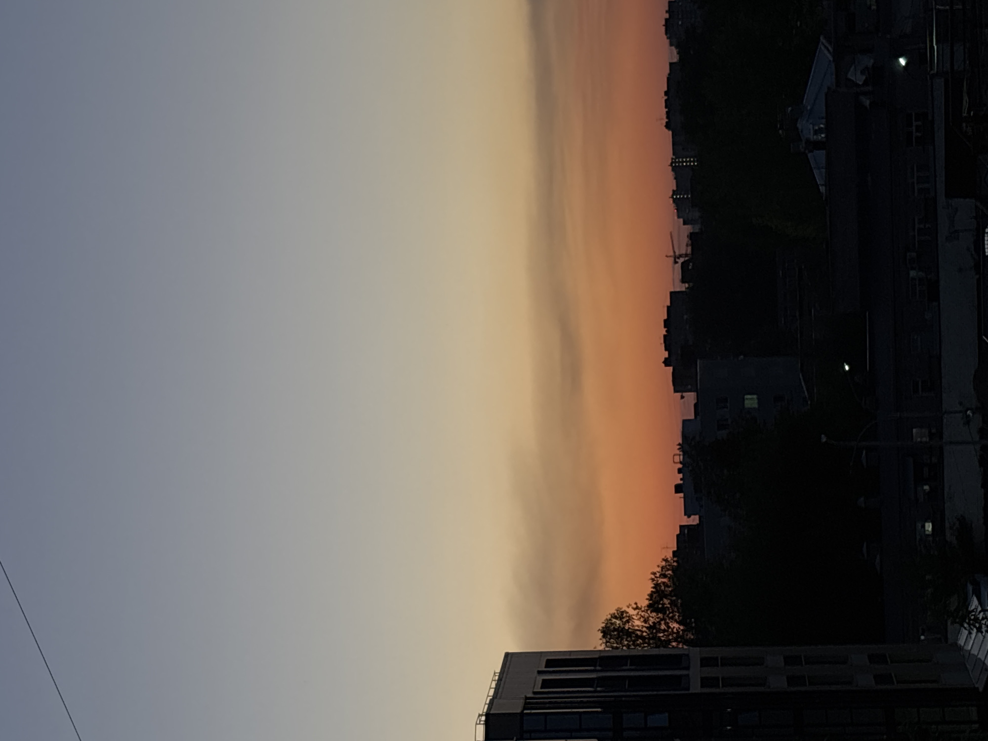 The image depicts a dusk or dawn urban scene, likely in Perm, Russia, as the prompt suggests. The sky transitions from a light greyish-blue to orange near the horizon, possibly indicating a sunset or sunrise. Silhouetted buildings and trees are visible against the lighter portion of the sky. A construction crane is also visible. The buildings are mostly dark, with a few lights visible. The scene is predominantly dark due to the low light conditions. A wire runs across the left portion of the sky. The image appears to have been taken from a relatively high vantage point.
