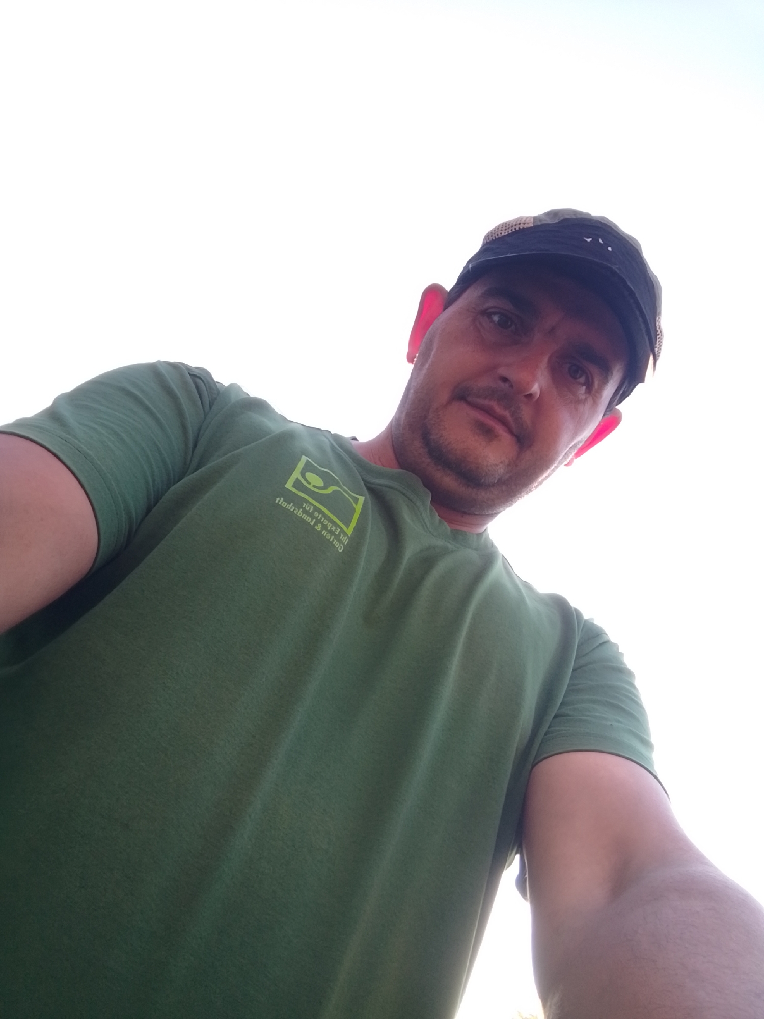 A man wearing a green t-shirt and a baseball cap is captured in a low-angle, selfie-style shot. The man's face is partially visible, with his eyes looking towards the camera. He has a light beard and is wearing a cap that partially obscures his forehead. The t-shirt has a small, light green logo on the left chest area. The background is predominantly a bright, overcast sky, suggesting an outdoor setting. The image's composition and lighting are consistent with a photograph taken during daylight hours. The location is the city of Wedel, Germany.