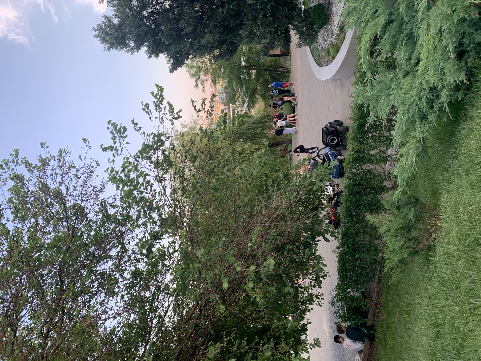 The media file shows an outdoor scene with a group of people gathered on a paved walkway. The walkway appears to be in a park or a public space, with lush greenery surrounding it, including trees and manicured bushes. In the background, there are buildings visible, suggesting an urban or suburban setting. A few electric scooters or similar personal mobility devices are parked near the walkway.
The people are dressed casually and appear to be socializing or engaged in some activity, possibly a group exercise or a casual gathering. Some individuals are standing, while others are seated on a low grassy area adjacent to the walkway.
The time of day appears to be late afternoon or early evening, as indicated by the soft, warm light and the sky's color, which has hints of pink and orange, suggesting sunset. The weather seems pleasant, with clear skies and ample daylight.
Notable details include the modern design of the walkway with a curved, white concrete border and a patch of decorative gravel. The overall atmosphere suggests a relaxed and communal gathering in a well-maintained outdoor environment. No visible text is present in the media file.