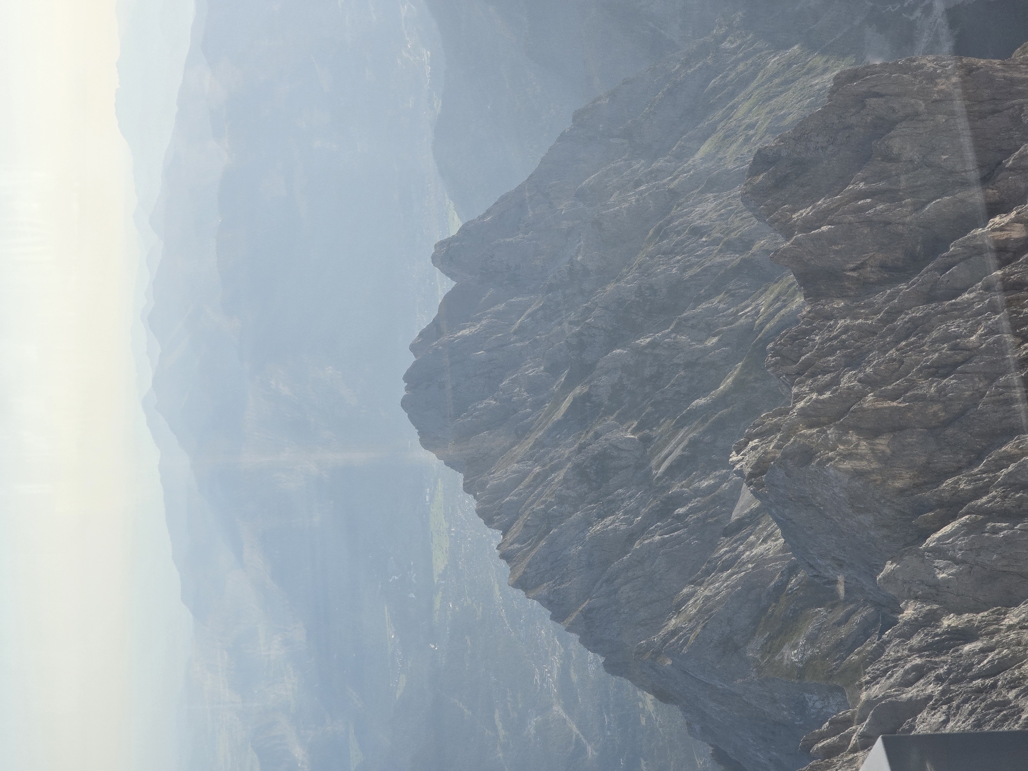 The image depicts a mountainous landscape, likely taken from a high vantage point. In the foreground, a rugged, rocky mountain ridge dominates the scene, with sharp inclines and visible strata. The texture of the rock is rough and weathered, with patches of sparse vegetation clinging to some slopes.
In the mid-ground, another, larger mountain peak rises, its slopes also characterized by sharp, angular formations and deep crevices. The surface of this mountain appears to be covered in scree and rock.
Further in the background, a series of progressively more distant mountain ranges fade into a hazy, atmospheric perspective. The furthest mountains are softened by atmospheric conditions, appearing as faint, undulating silhouettes against a pale sky.
Below the main mountain formations, a valley or plain is visible, dotted with what appear to be buildings and fields, suggesting human settlement. The color palette is muted, dominated by grays, blues, and greens, with the sky being a pale, hazy white. The overall impression is one of immense scale and natural grandeur, possibly a view of the Bavarian Alps. The presence of a faint rainbow-like refraction on the right side of the image suggests it may have been taken through glass, potentially from a cable car or observation deck. Without further information, pinpointing the exact location in Grainau, Germany, is not possible from the image alone, but the visual cues are consistent with the topography of that region.