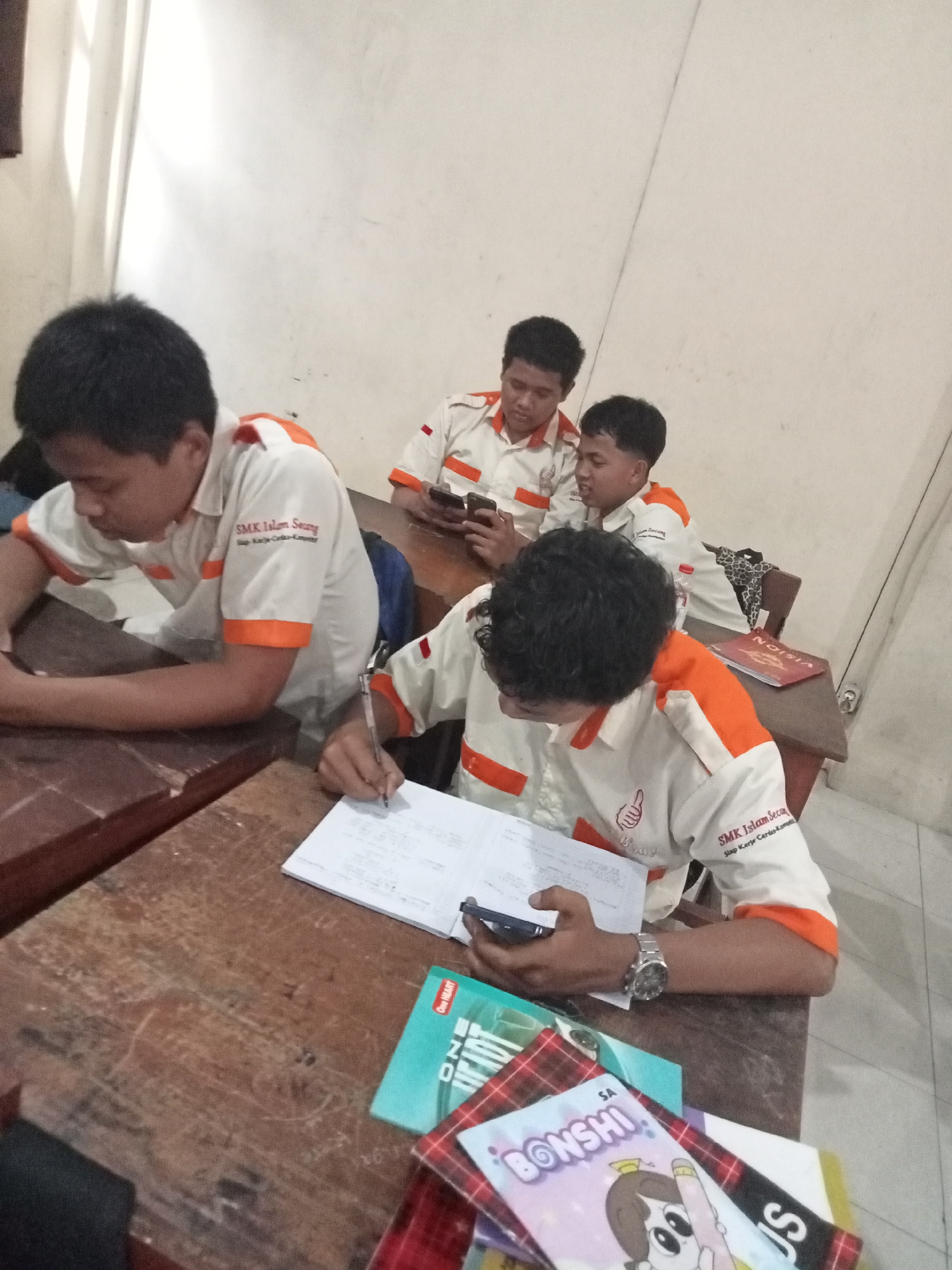 The image depicts a classroom scene in Secang, Indonesia, likely at SMK Islam Secang, as evidenced by the school emblem on the students' uniforms. Four young male students, all wearing similar white polo shirts with orange trim and the school logo, are seated at wooden desks. Two students in the foreground are engaged in activities at their desks. The student on the left, facing away from the camera, appears to be looking down at something on his desk. The student in the center foreground, also with his back to the camera, is actively writing in a notebook with a pen. He is wearing a watch on his left wrist and holding a mobile phone in his right hand. In the background, two other students are seated at their desks. The student on the left in the background is looking at a mobile phone, while the student on the right in the background is also looking at a mobile phone. The classroom environment is simple, with light-colored walls and wooden desks. Several books and notebooks are visible on the desks, suggesting an academic setting. The lighting appears to be artificial, possibly from overhead lights.